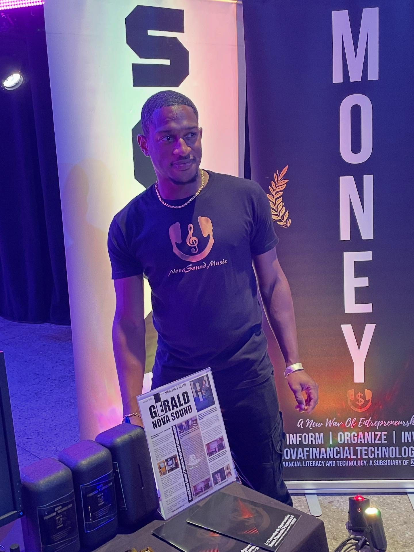 Young man standing next to a display with posters and banners, wearing a black T-shirt with a logo, in an indoor event space.