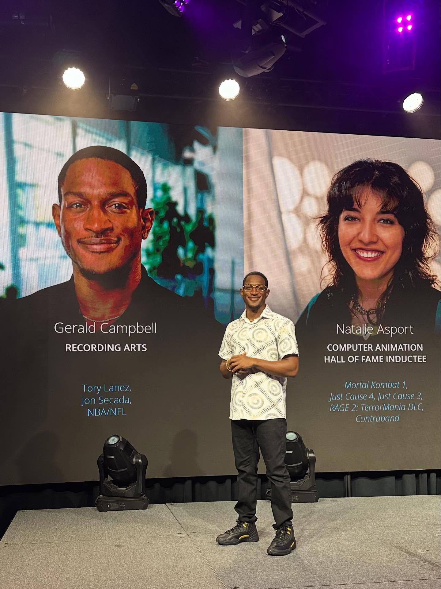 Man standing on stage with large screen behind showing two people, names Gerald Campbell and Natalie Asport, with descriptions.