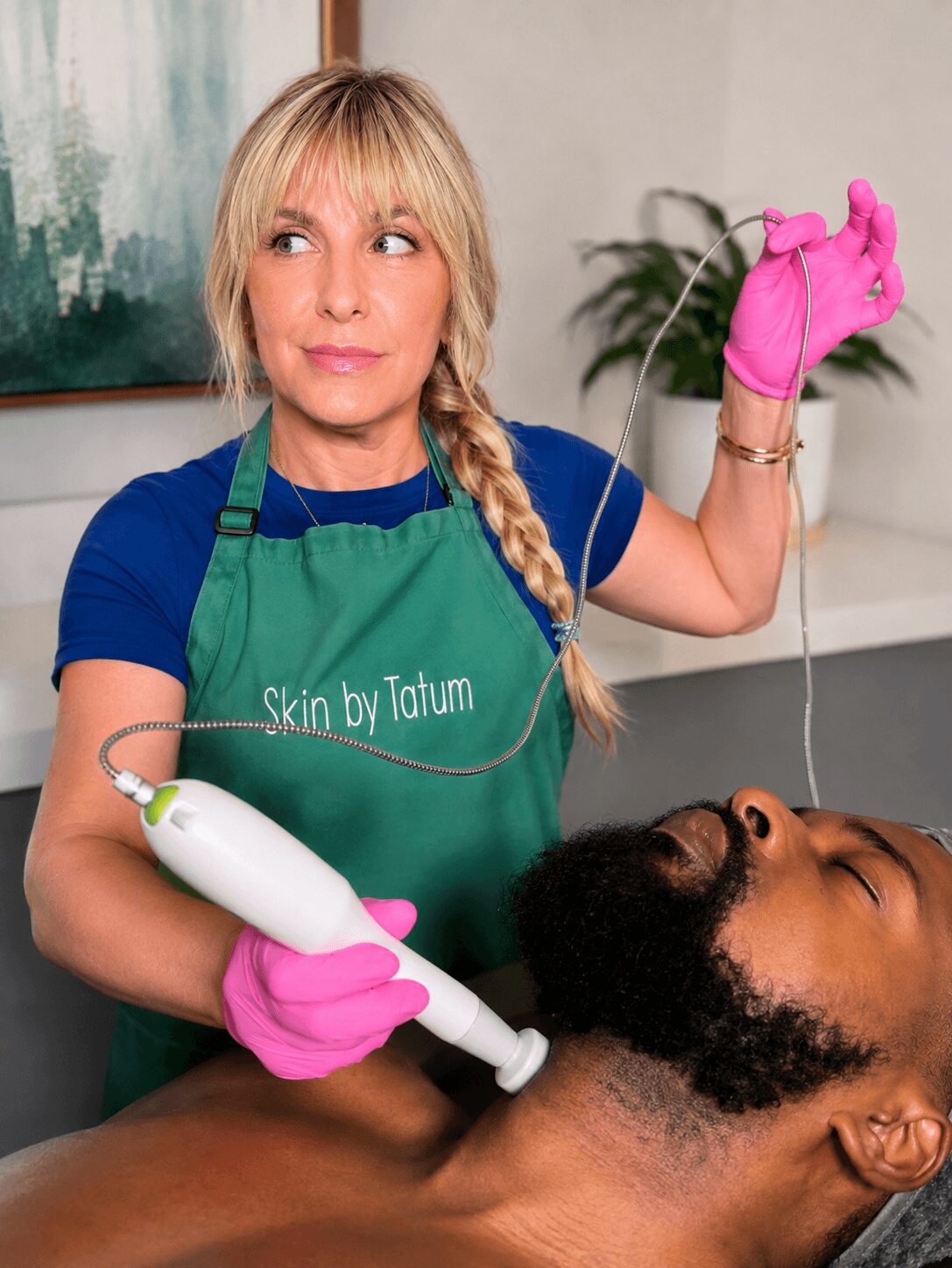 Woman in green apron and pink gloves performs a facial treatment on a man with a device, in a spa setting.