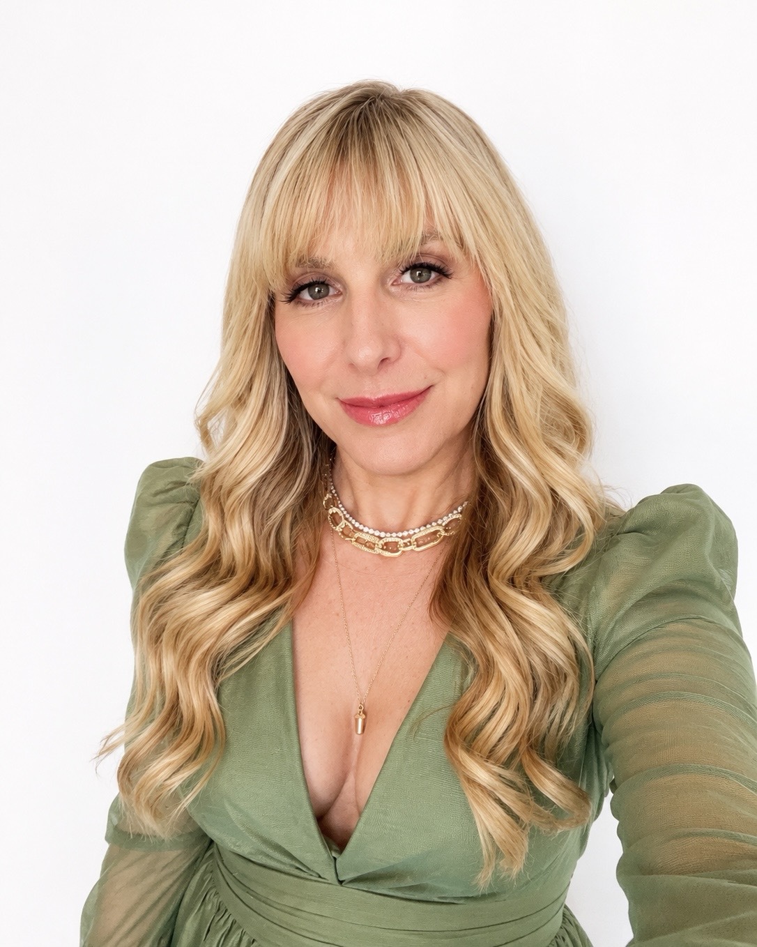 Woman with long blonde hair wearing a green top and layered necklaces, smiling at the camera.