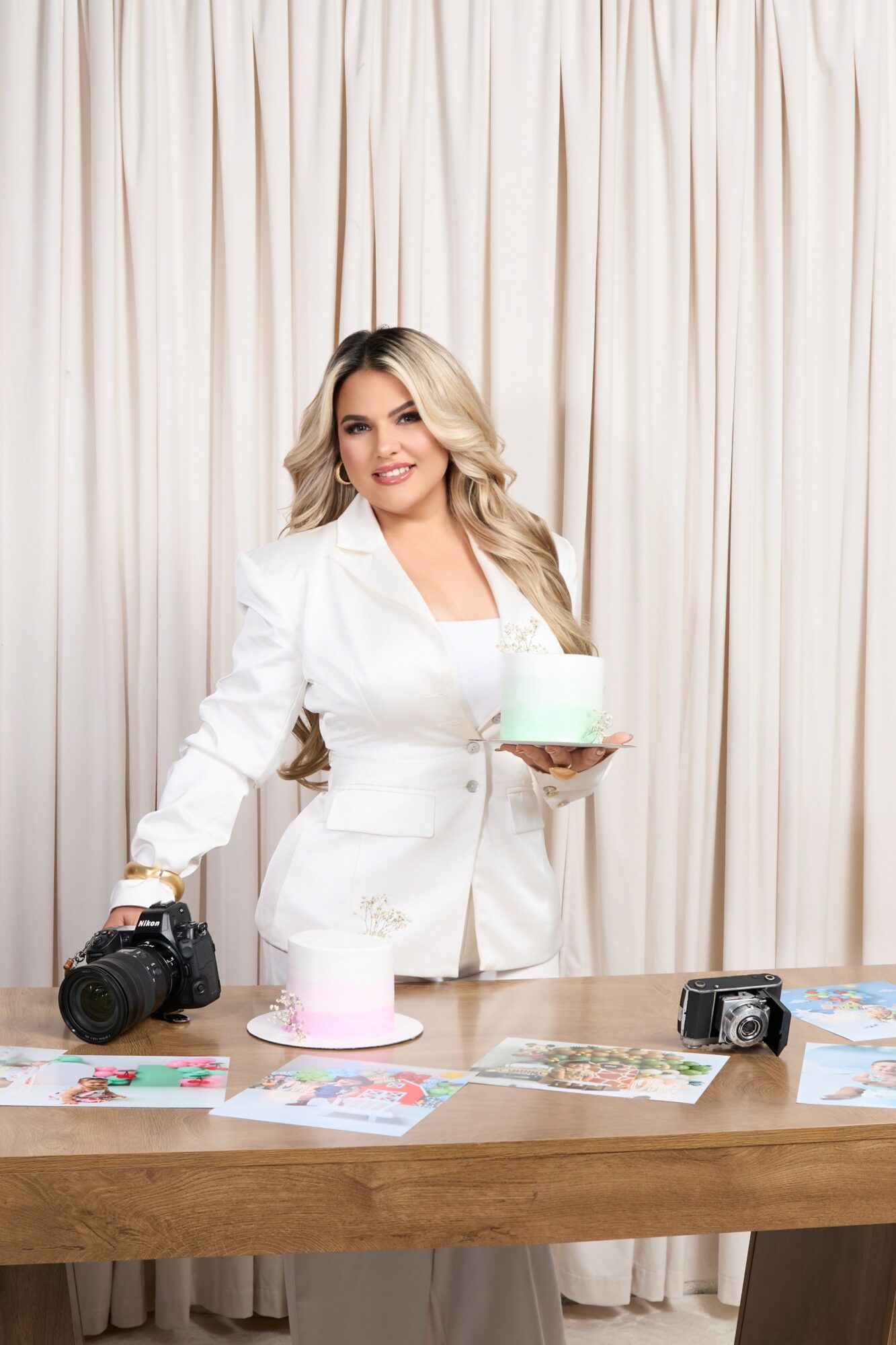 Woman in white blazer holding a cake, standing behind a table with cameras and papers, cream-colored curtain background.