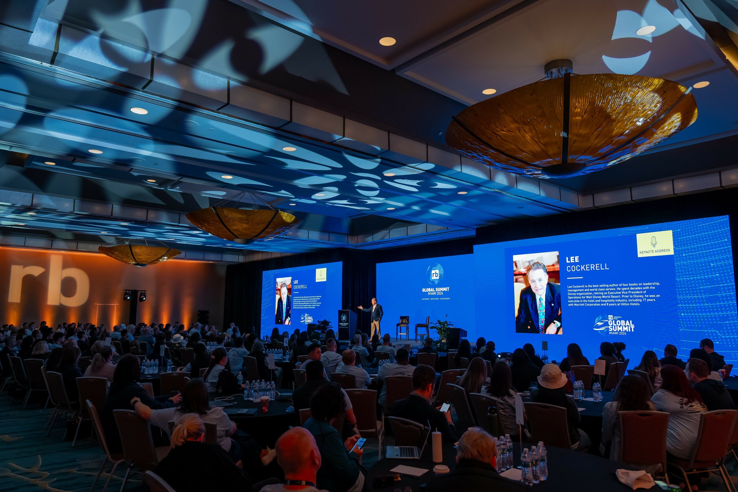 Large conference room filled with seated audience watching a presentation on stage with multiple screens, ceiling lights, and decorative ceiling patterns.