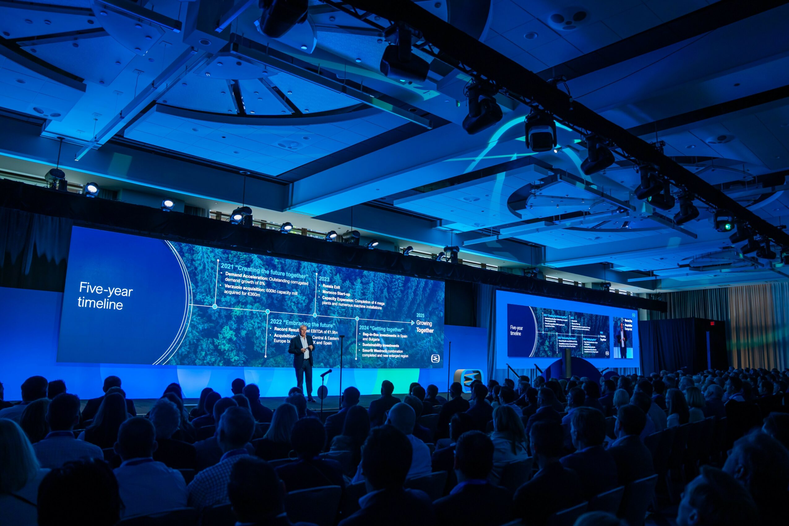 A presenter stands on stage in front of a large screen displaying a timeline and text, with an audience watching attentively.