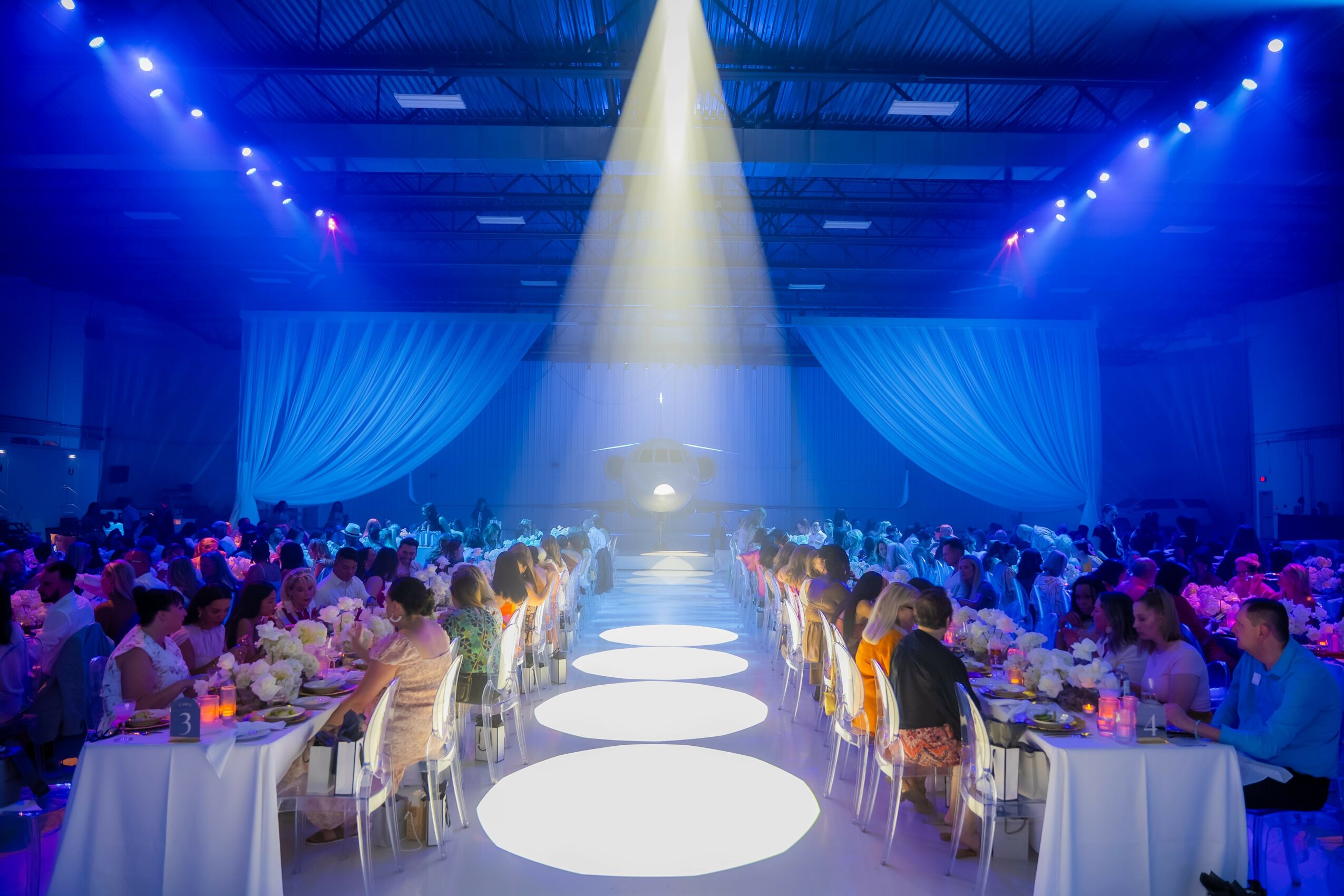 Large event hall with round tables, people dining, stage with bright spotlight, blue lighting, and draped curtains.