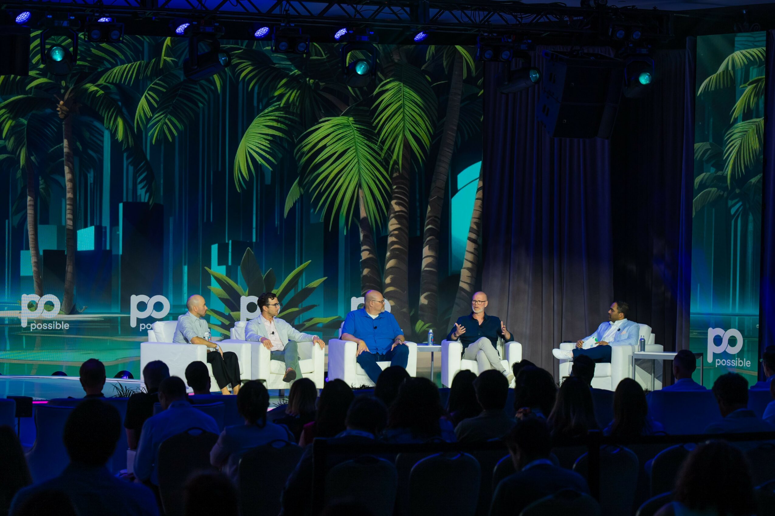 Panel discussion with five people on stage, audience in foreground, tropical-themed backdrop with palm trees, blue lighting.