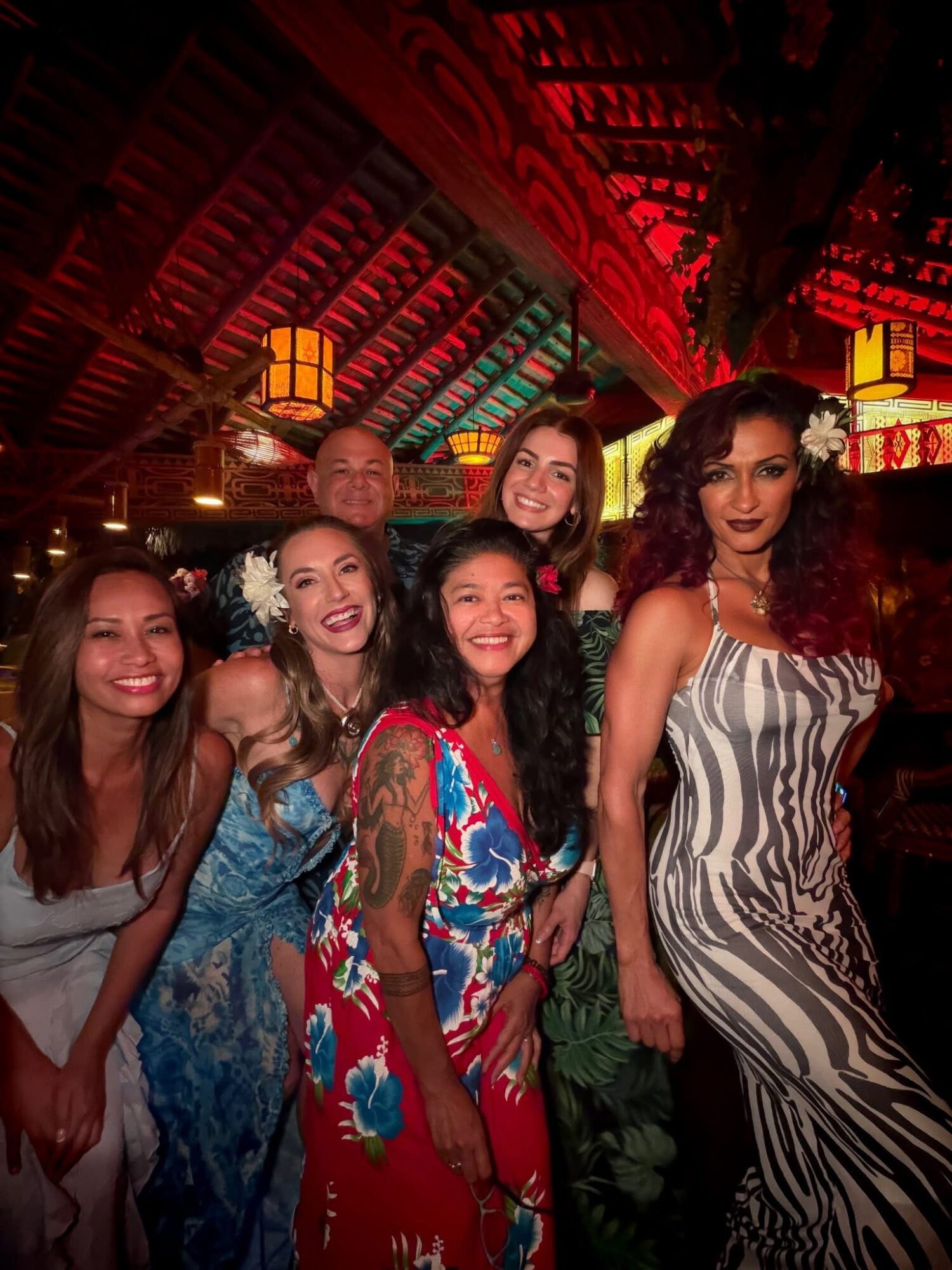 Six women smiling and posing together indoors with colorful lighting and a wooden ceiling.