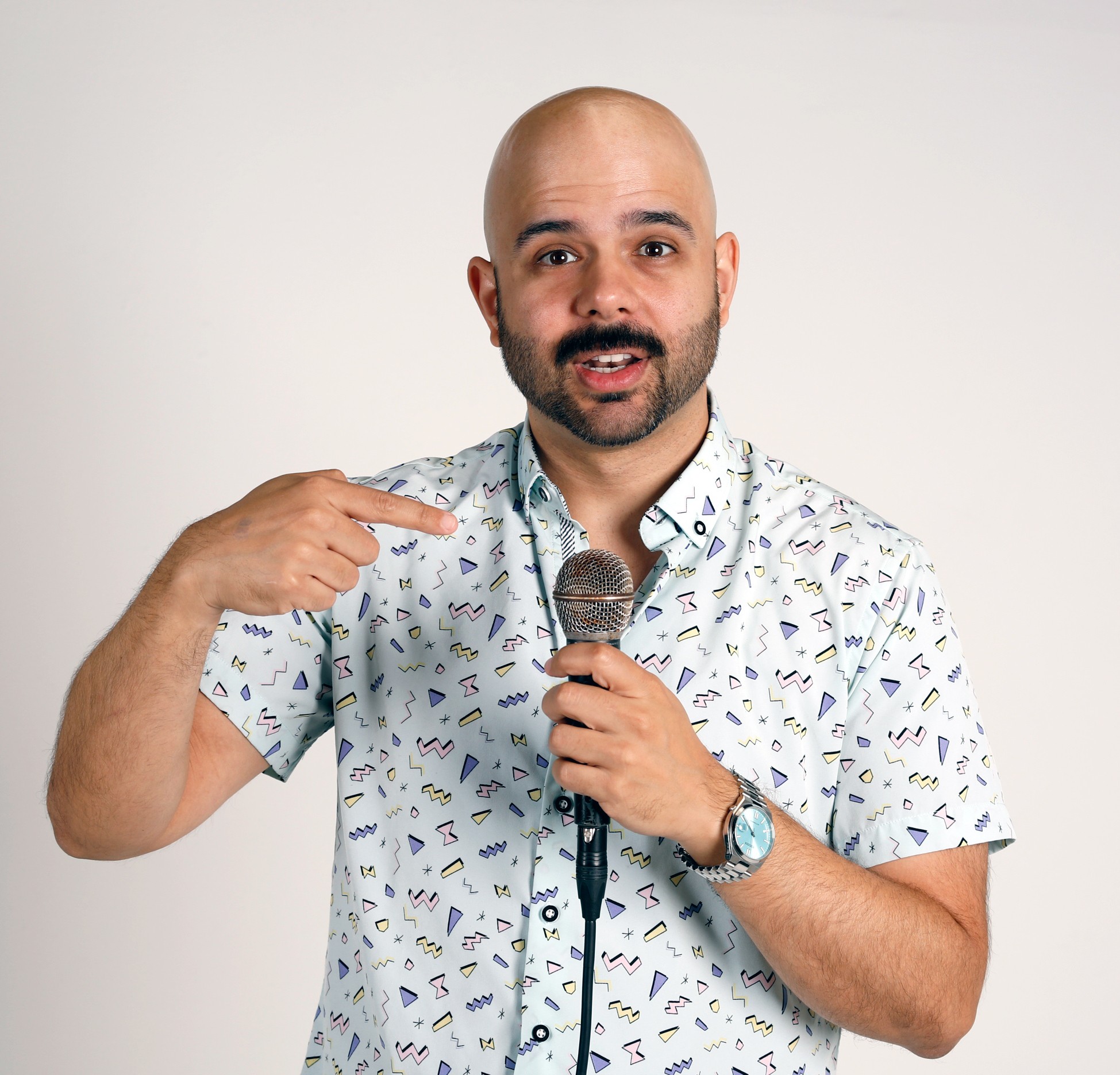 Bald man with beard holding microphone, pointing to his head, wearing a patterned short-sleeve shirt, standing against a plain background.