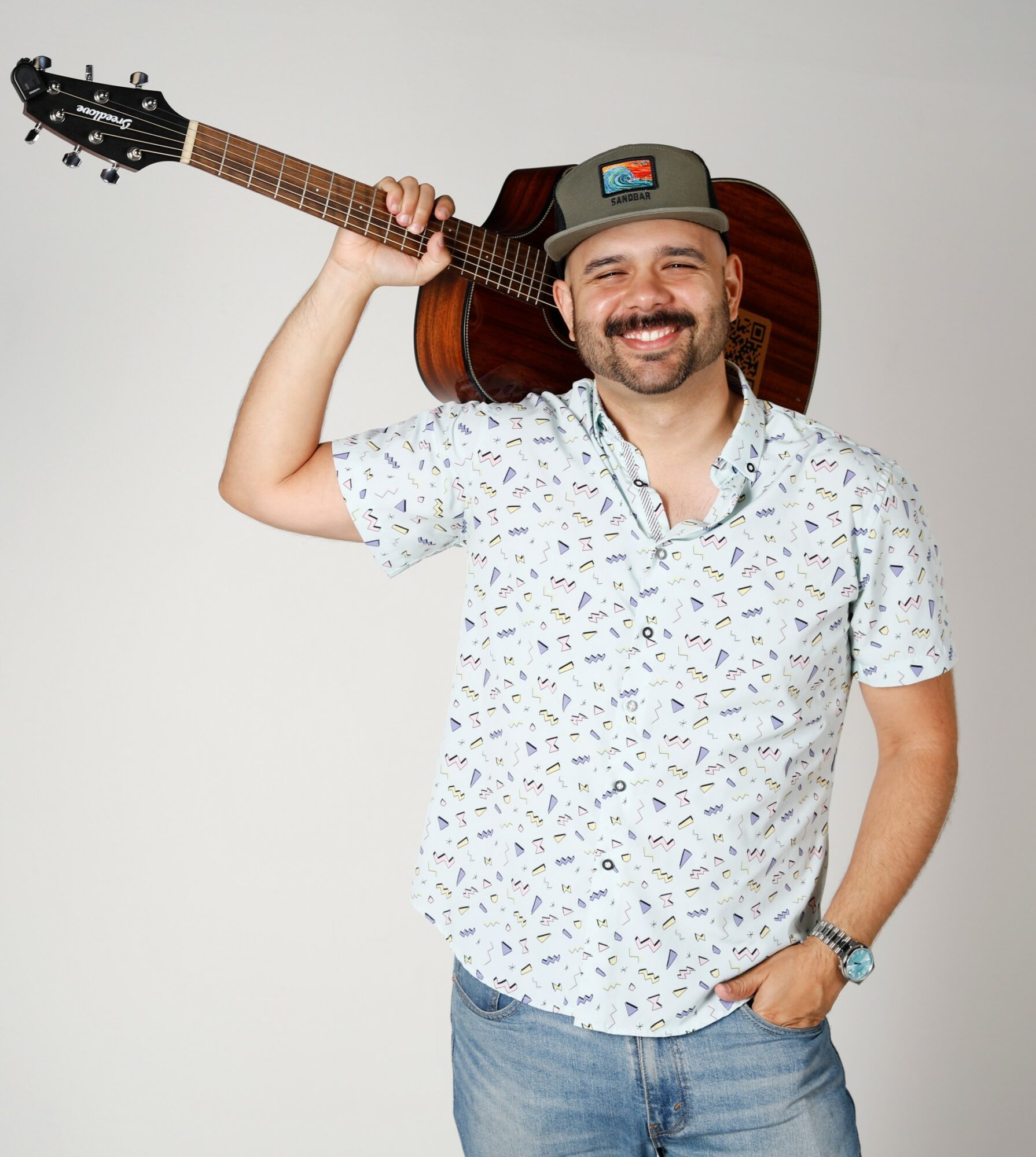Smiling man with a guitar on his shoulder, wearing a patterned shirt and a cap, standing against a plain background.