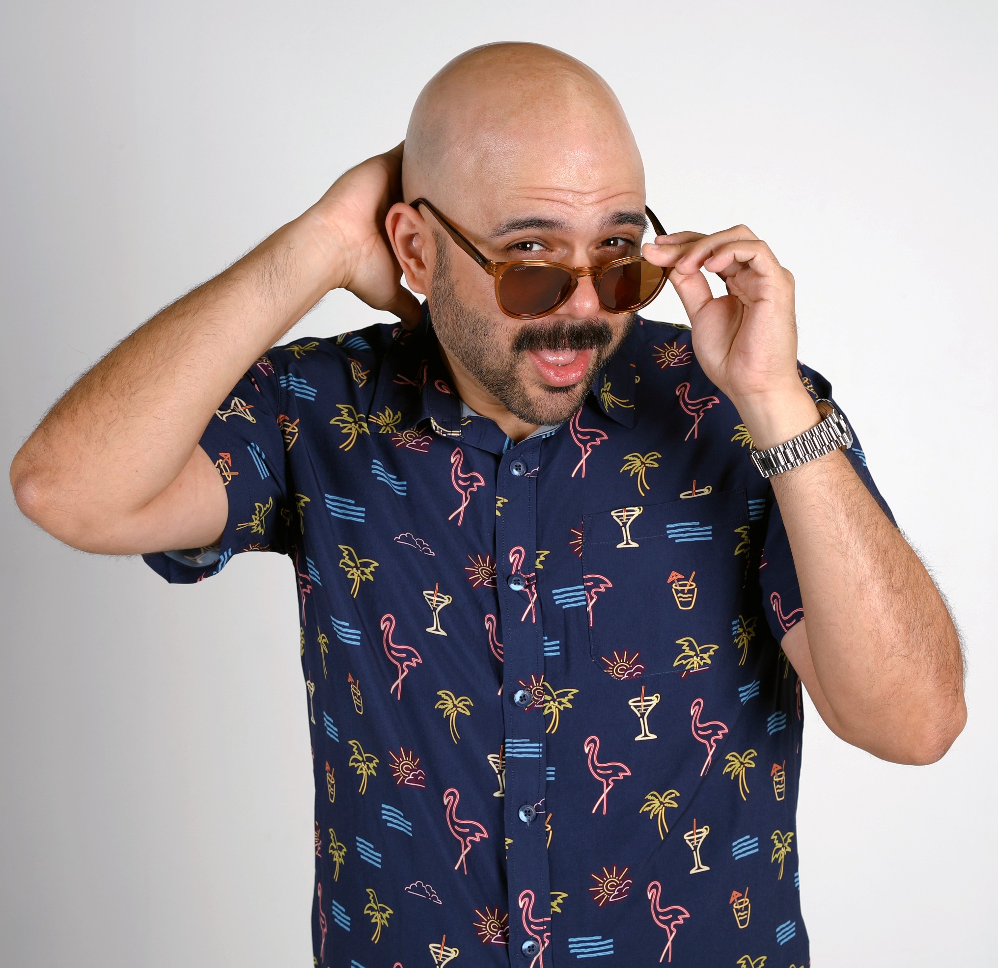 Bald man with sunglasses adjusting them, wearing a colorful tropical shirt, against a plain background.