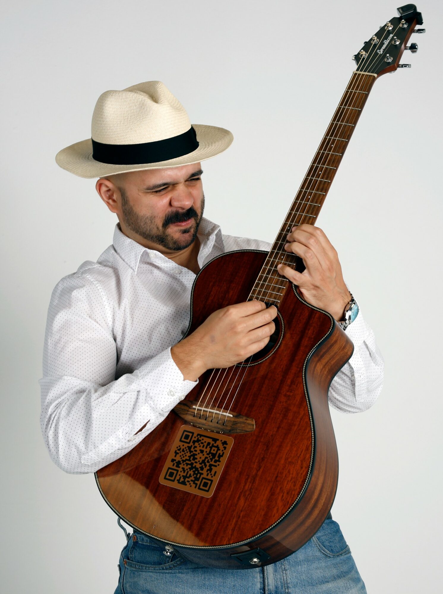Man wearing a white shirt and hat playing an acoustic guitar against a plain background.