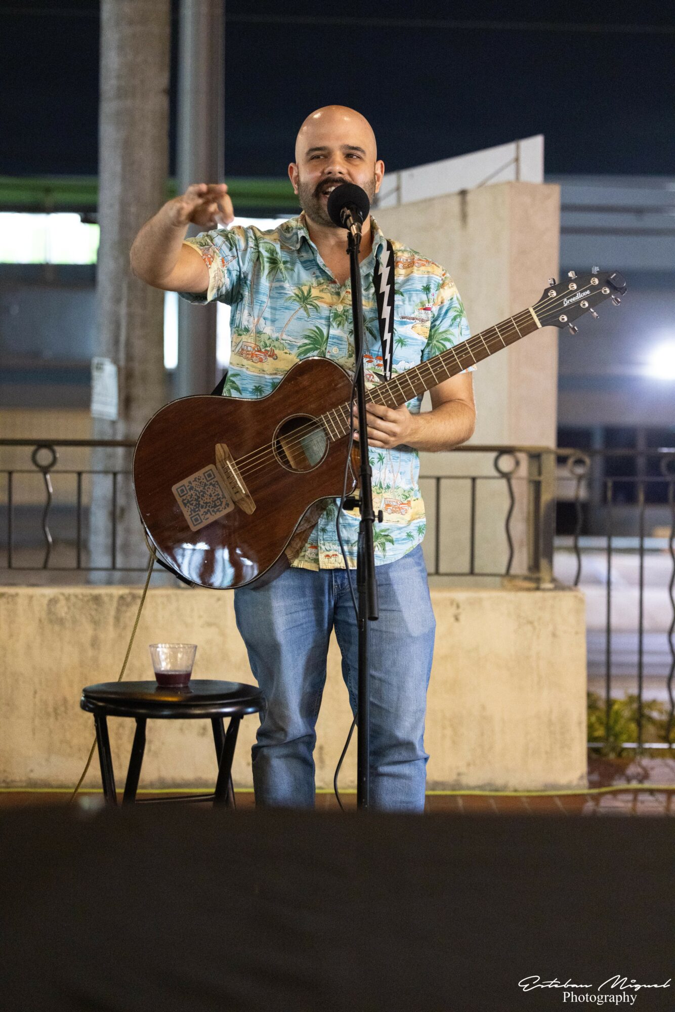 Man with a guitar performs on stage, gesturing with his right hand, with a microphone and a drink on a small table nearby.