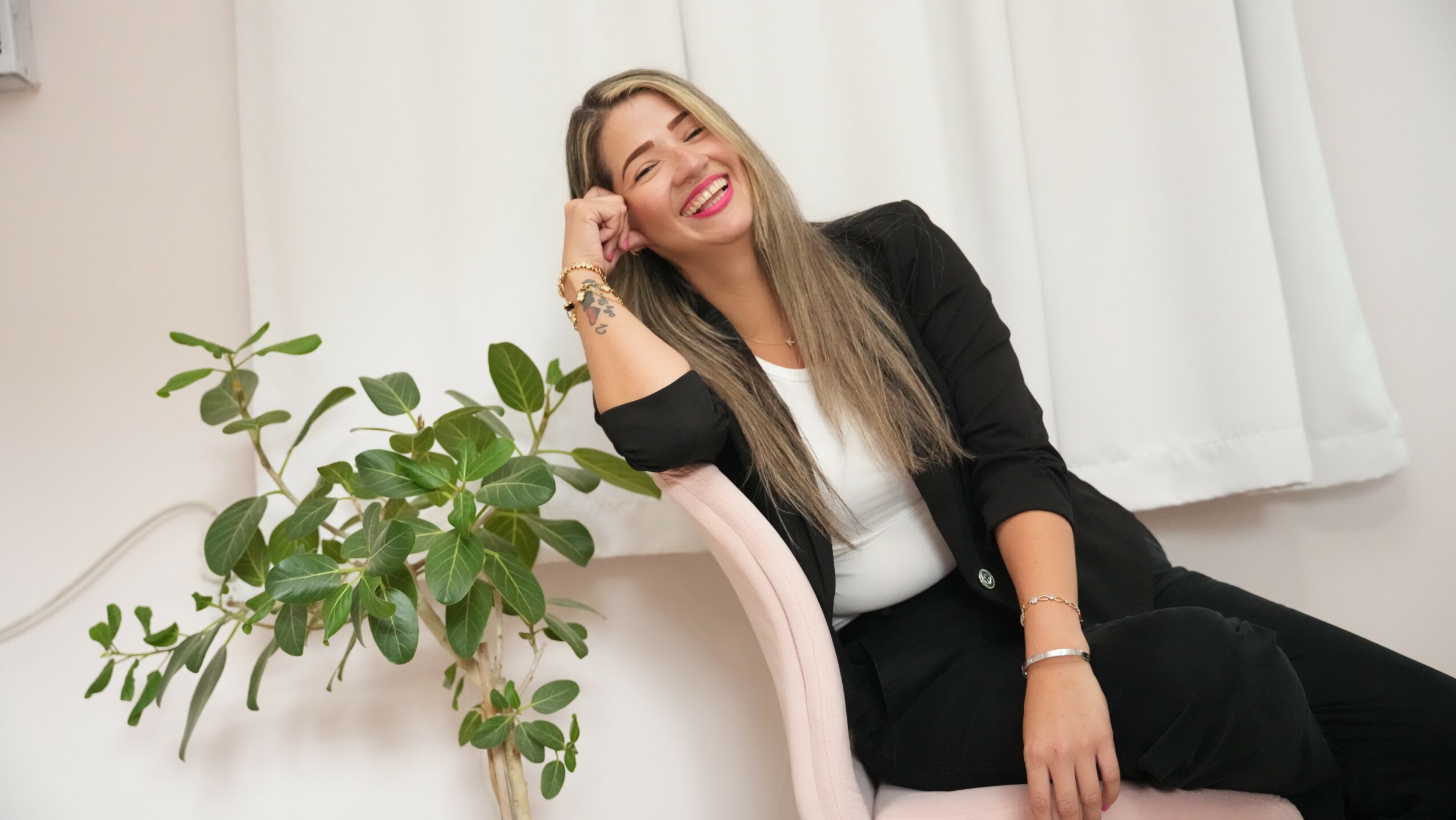 Woman with long hair smiling, sitting on a chair, wearing a black blazer and white top, next to a green plant.