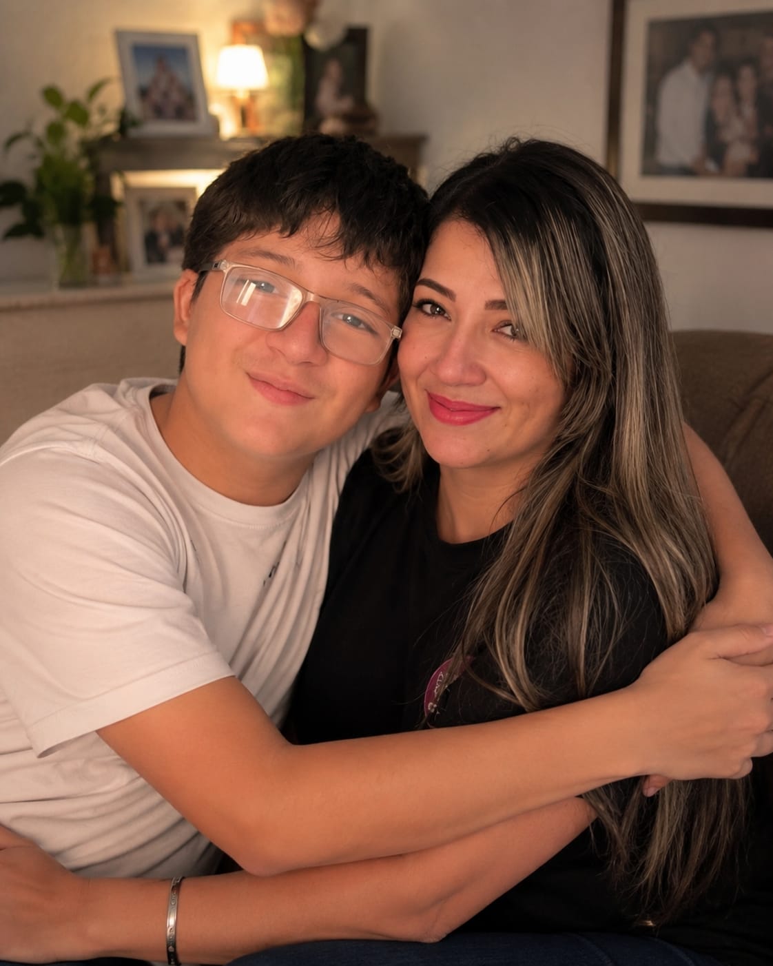 A young person and an adult woman hugging and smiling indoors, with framed photos and a lamp in the background.