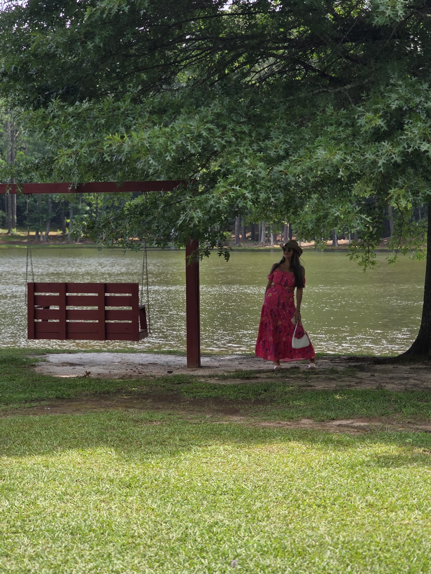 Person in a pink dress standing near a tree by a body of water, with a grassy area in foreground.