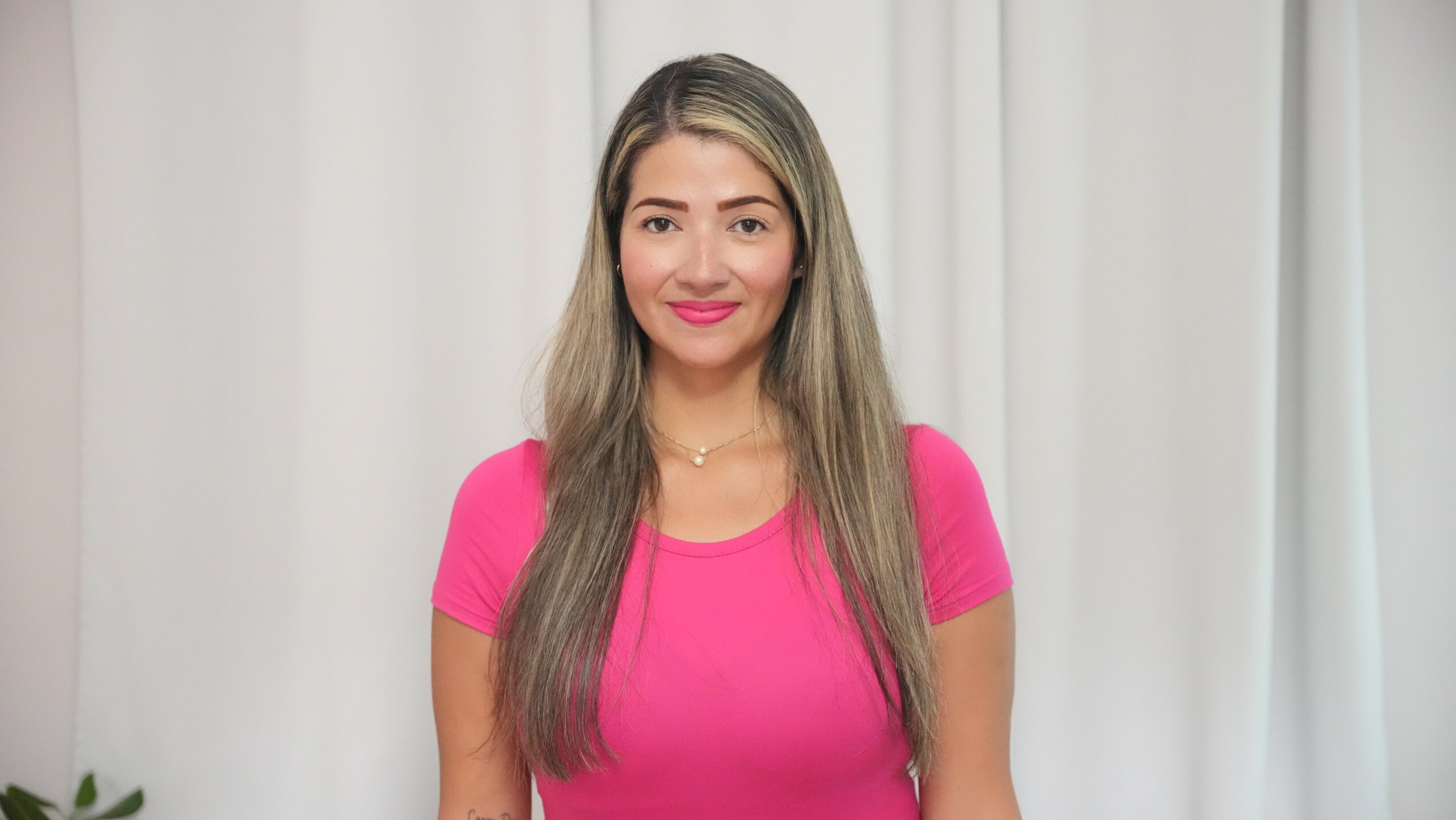 Woman with long hair wearing a pink shirt, smiling, standing in front of a white curtain backdrop.
