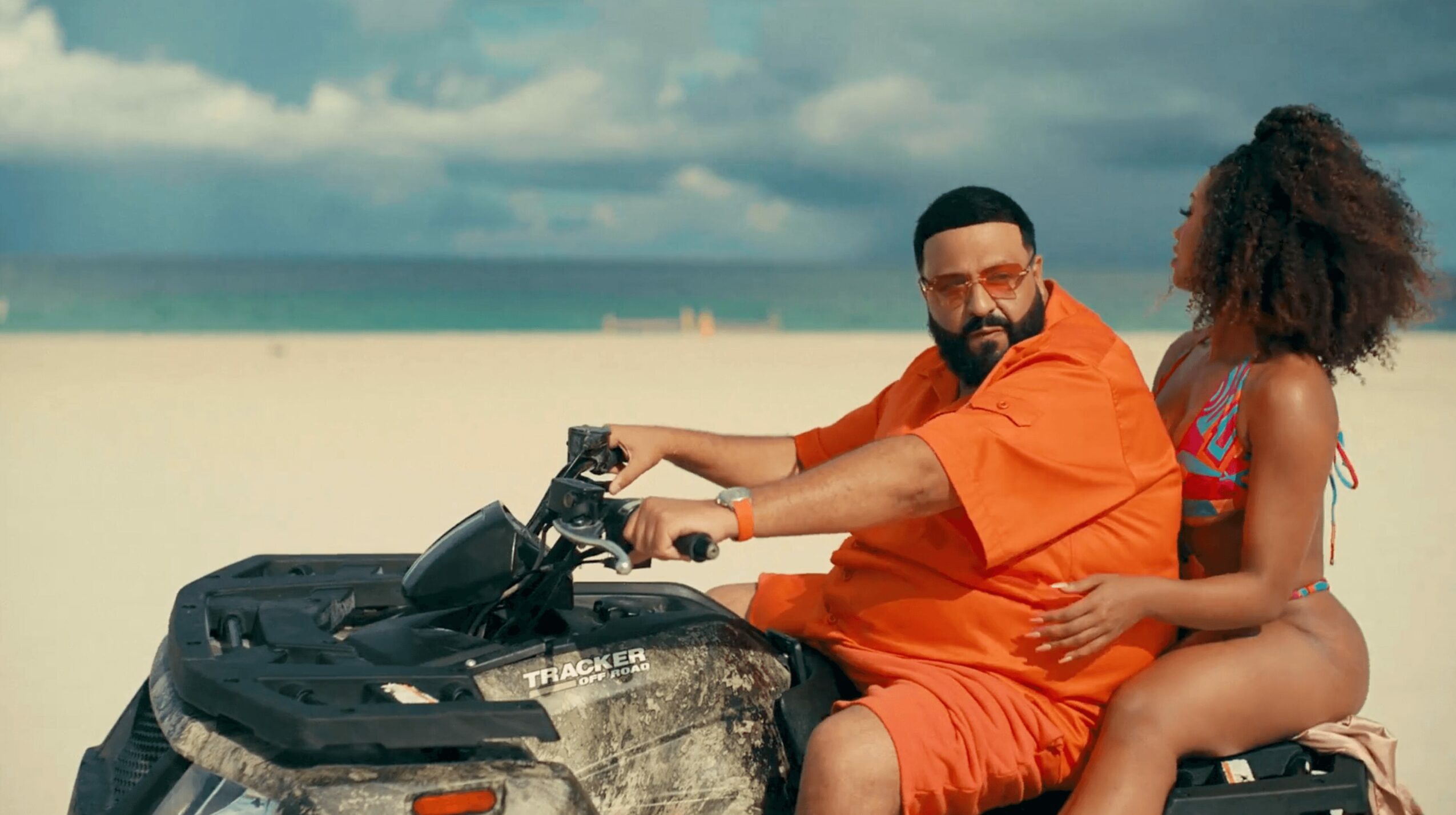 Two people sitting on a motorcycle on a beach, with ocean and sky in the background.