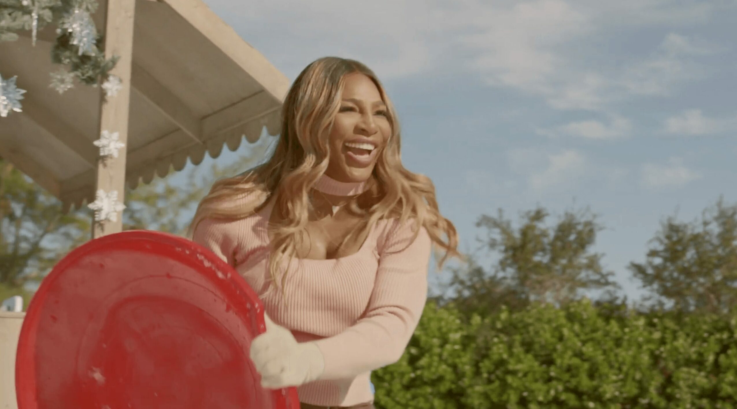Woman smiling outdoors holding a red object, with trees and a blue sky in the background.
