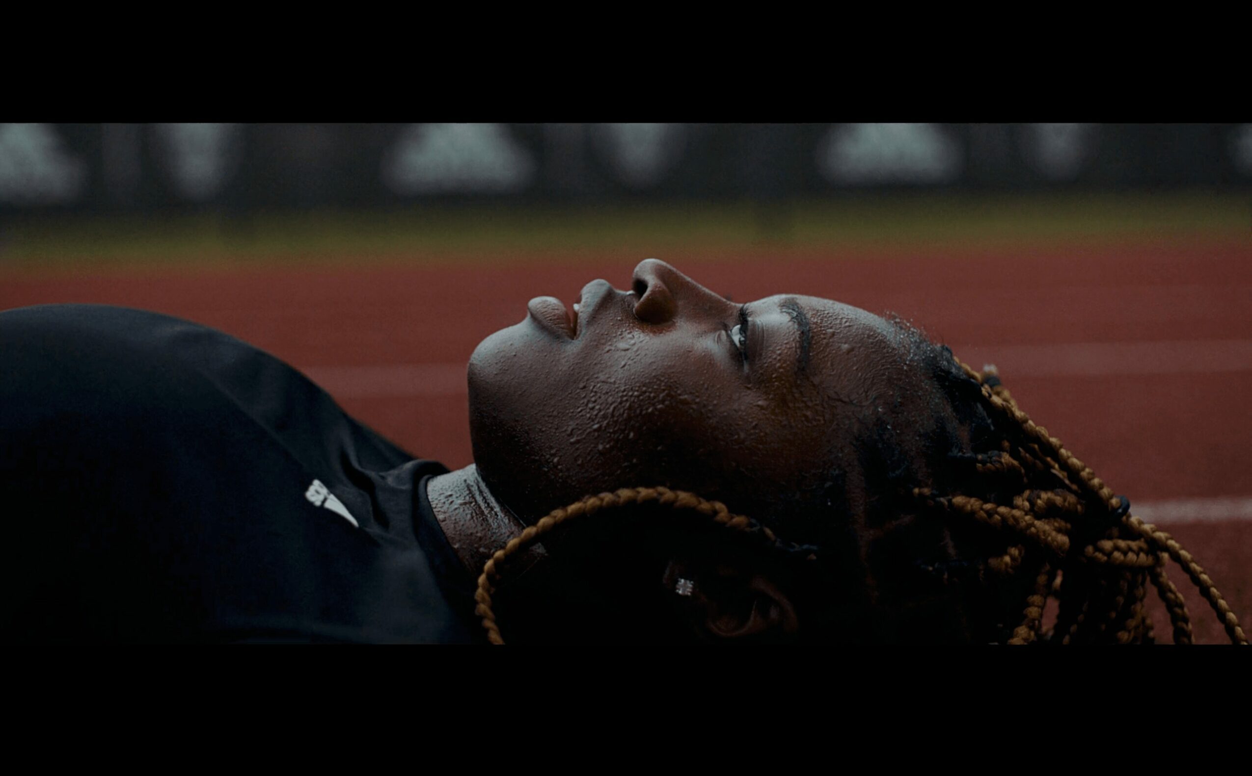 Person lying on a track with eyes closed, head tilted back, and braided hair visible.