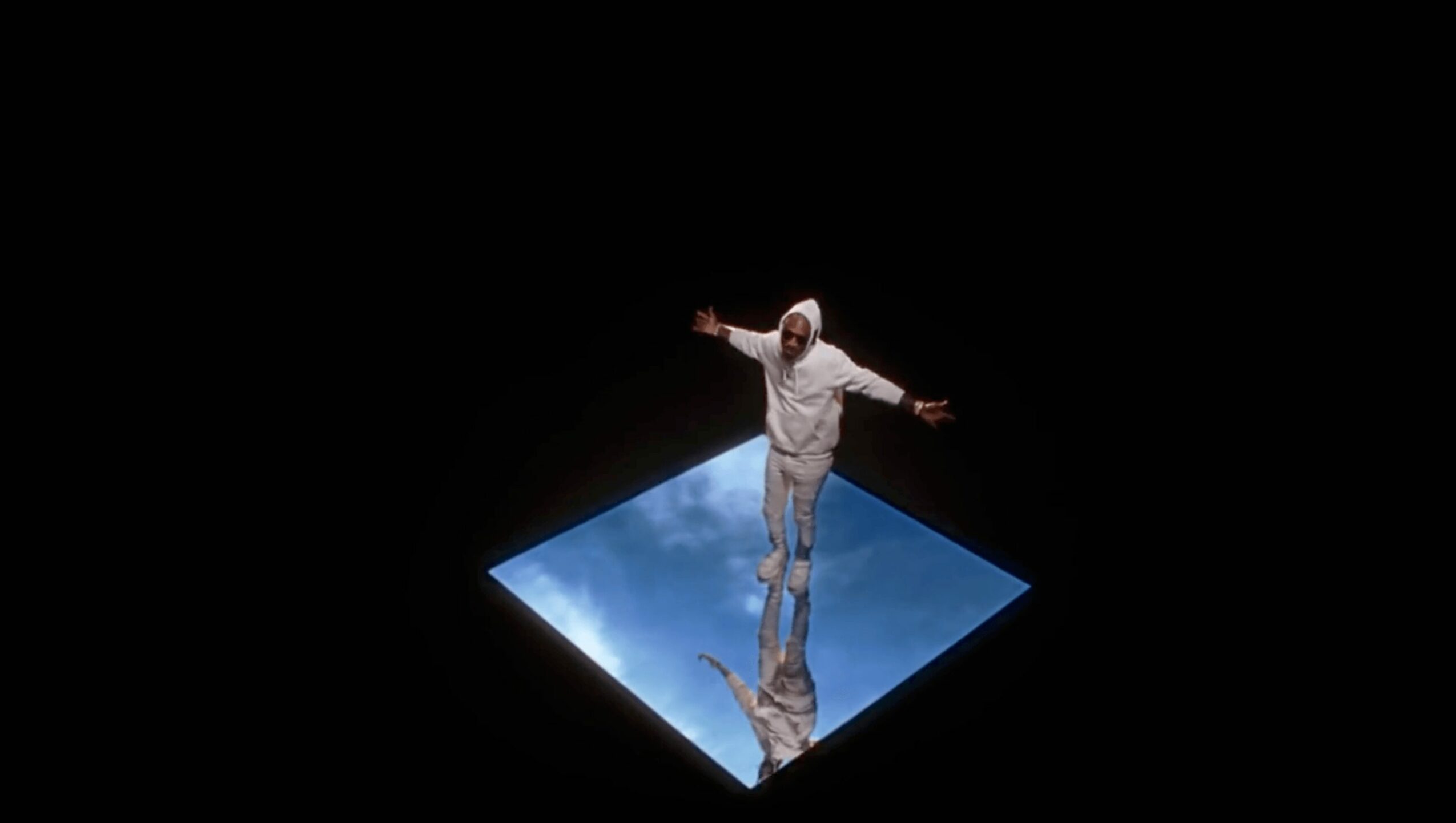 Person standing on a reflective surface with a blue sky and clouds, arms outstretched, against a black background.