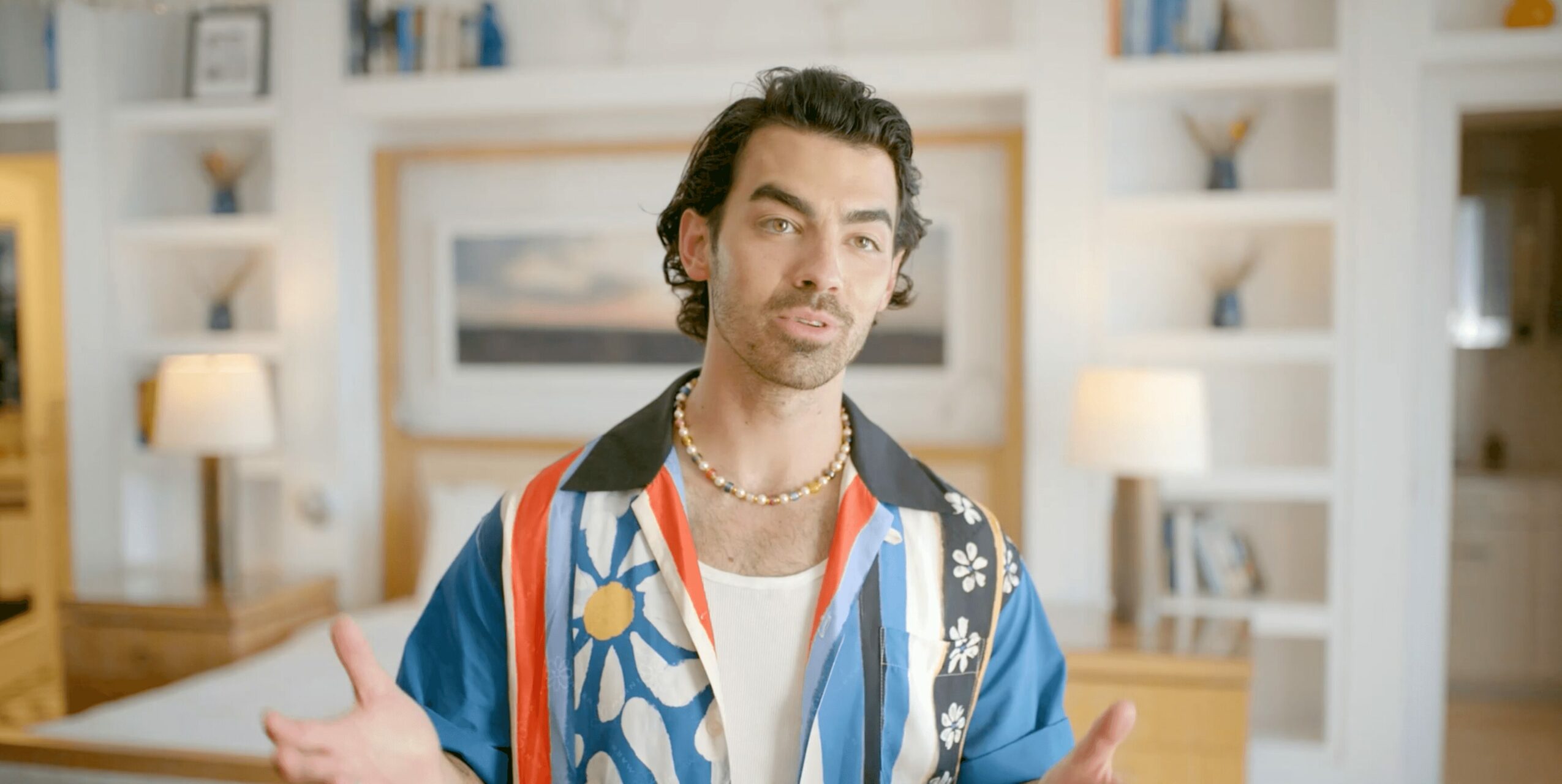 Young man with dark curly hair and a patterned shirt stands in a brightly lit room with shelves and lamps.