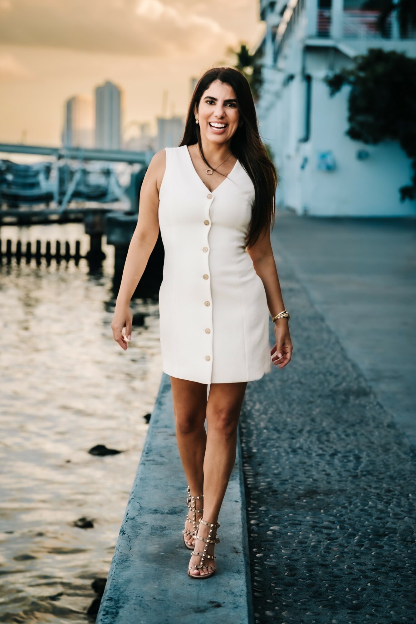 Woman in white dress walking on a pier with water and buildings in the background, smiling.