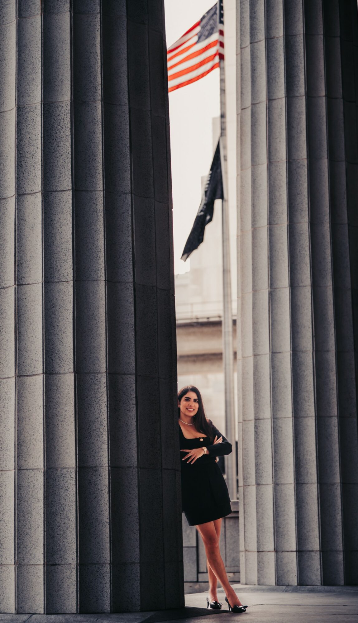 Woman in black dress standing between large gray columns with American flag visible in the background.