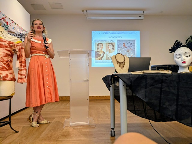 Woman in orange dress giving a presentation in a room with jewelry images on a screen, table with jewelry display, and mannequin head.