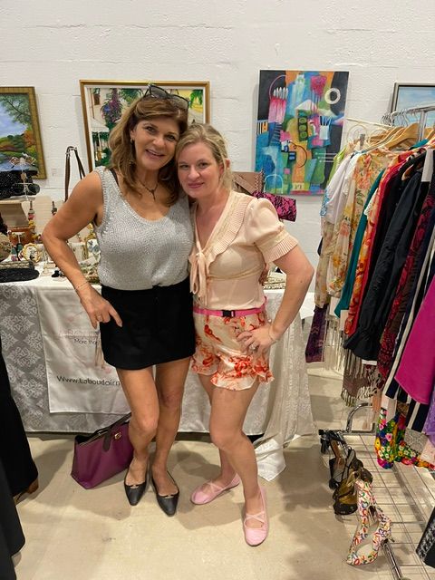 Two women standing in an art gallery, smiling, with colorful paintings on the wall behind them.