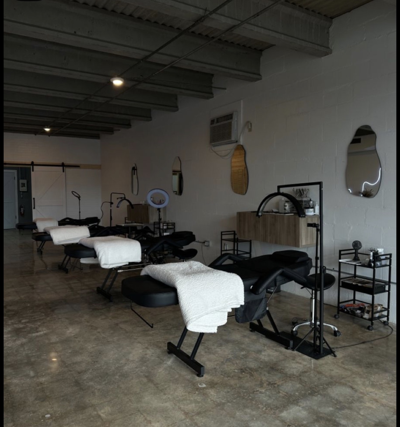 Salon with massage beds, towels, mirrors, and equipment, in a spacious room with a concrete floor and ceiling lights.