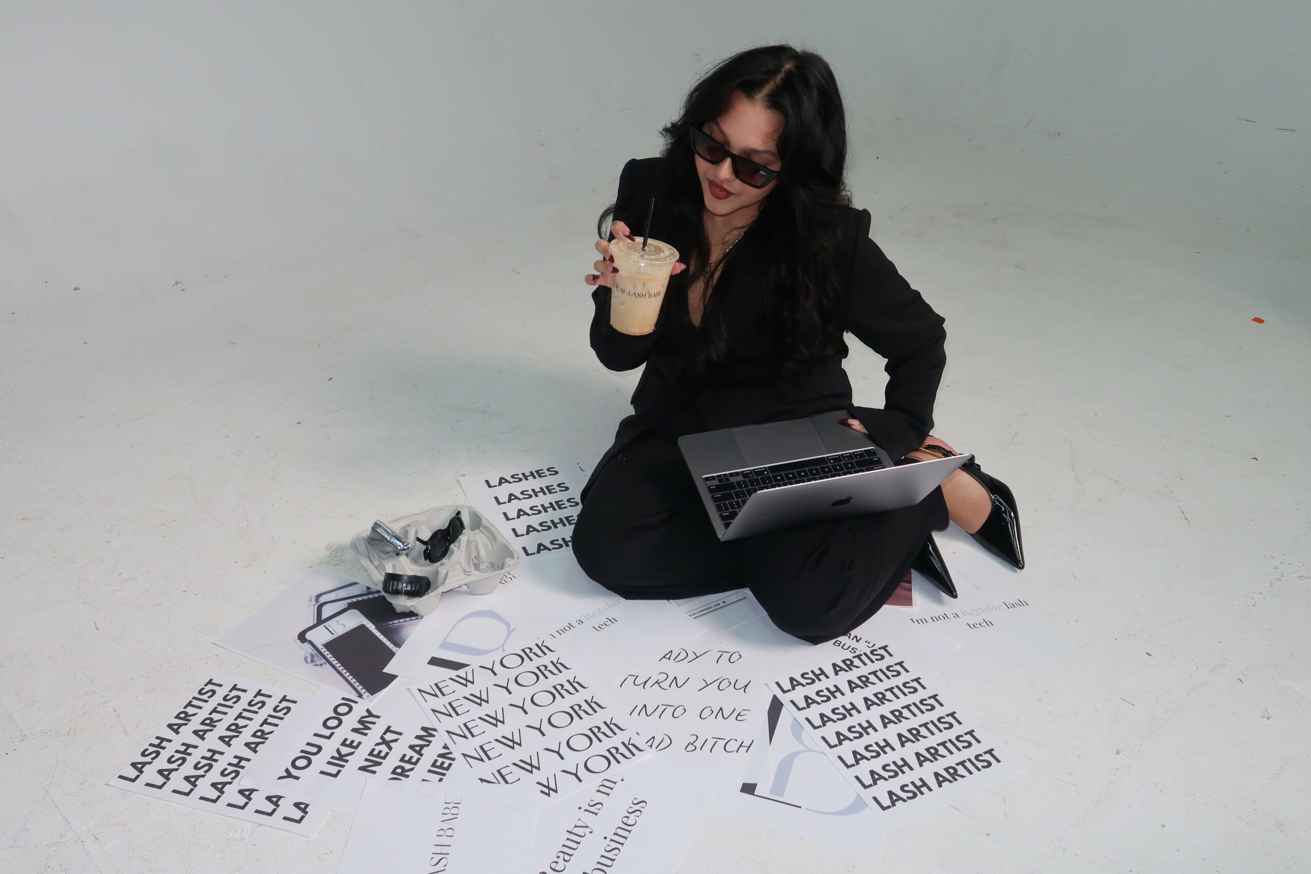 Woman sitting on the floor with a laptop, holding a drink, surrounded by printed papers, against a plain background.