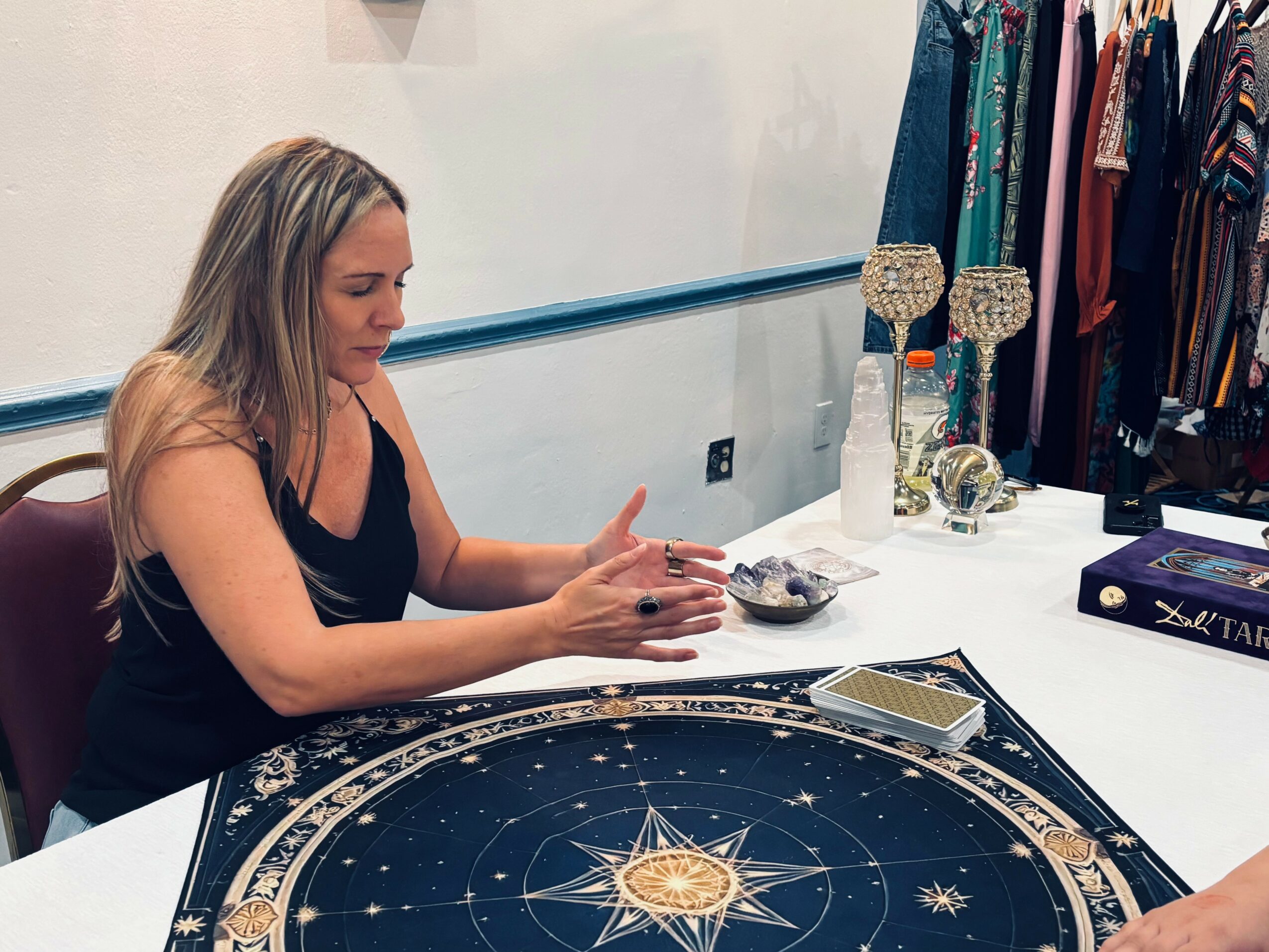 Woman sitting at a table with a large celestial-themed game board, surrounded by clothing racks and decorative items.
