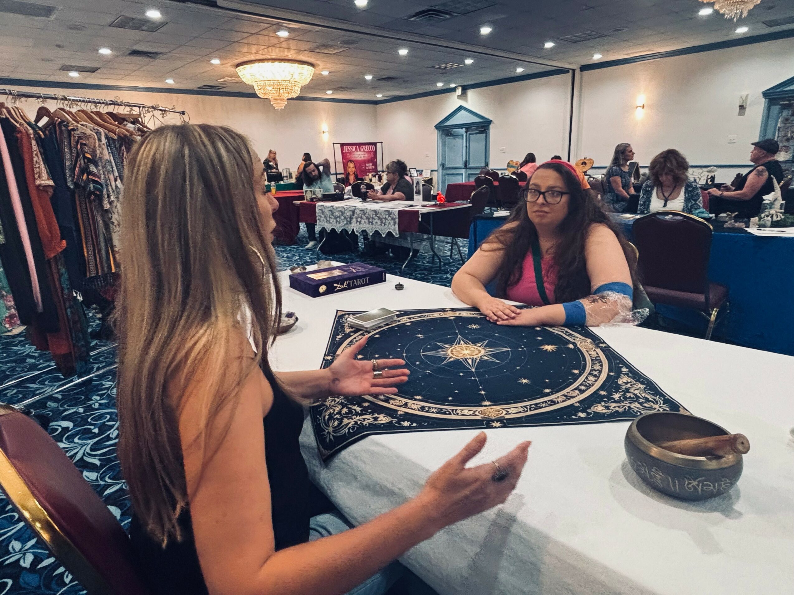 Two women sit at a table in a large, decorated room with other people and tables in the background.