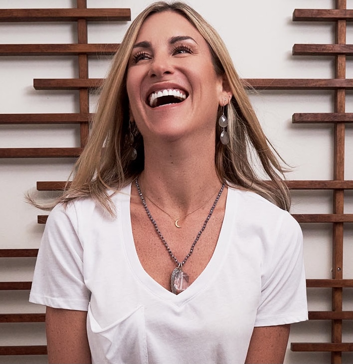 Woman with long blonde hair smiling, wearing a white top and layered necklaces, standing in front of a wooden wall.