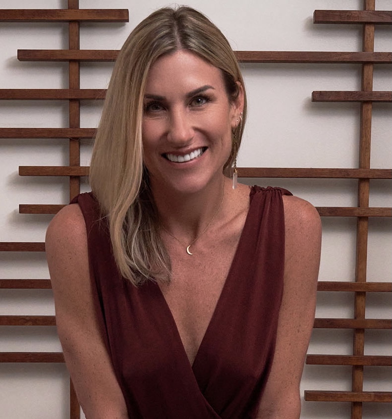 Woman with shoulder-length blonde hair smiling, wearing a sleeveless maroon top, standing in front of a wooden grid background.