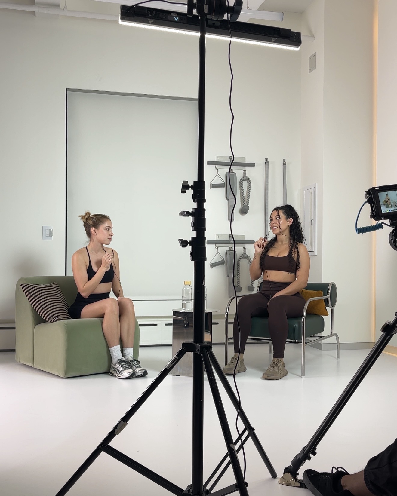 Two women sit and talk in a studio with equipment and a white wall, one on a green couch, the other on a chair.