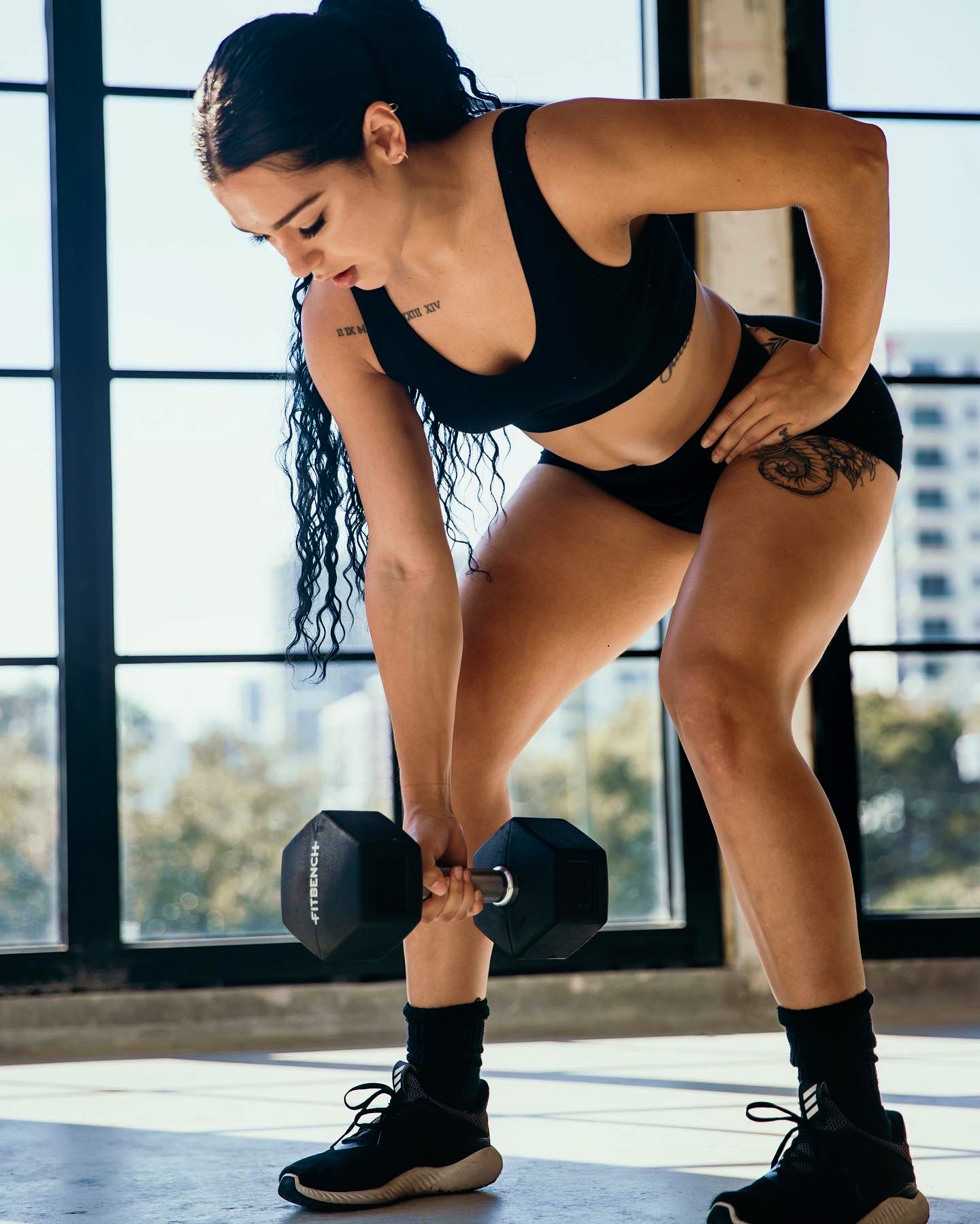 Woman in workout clothes lifting a dumbbell in a gym with large windows behind her.