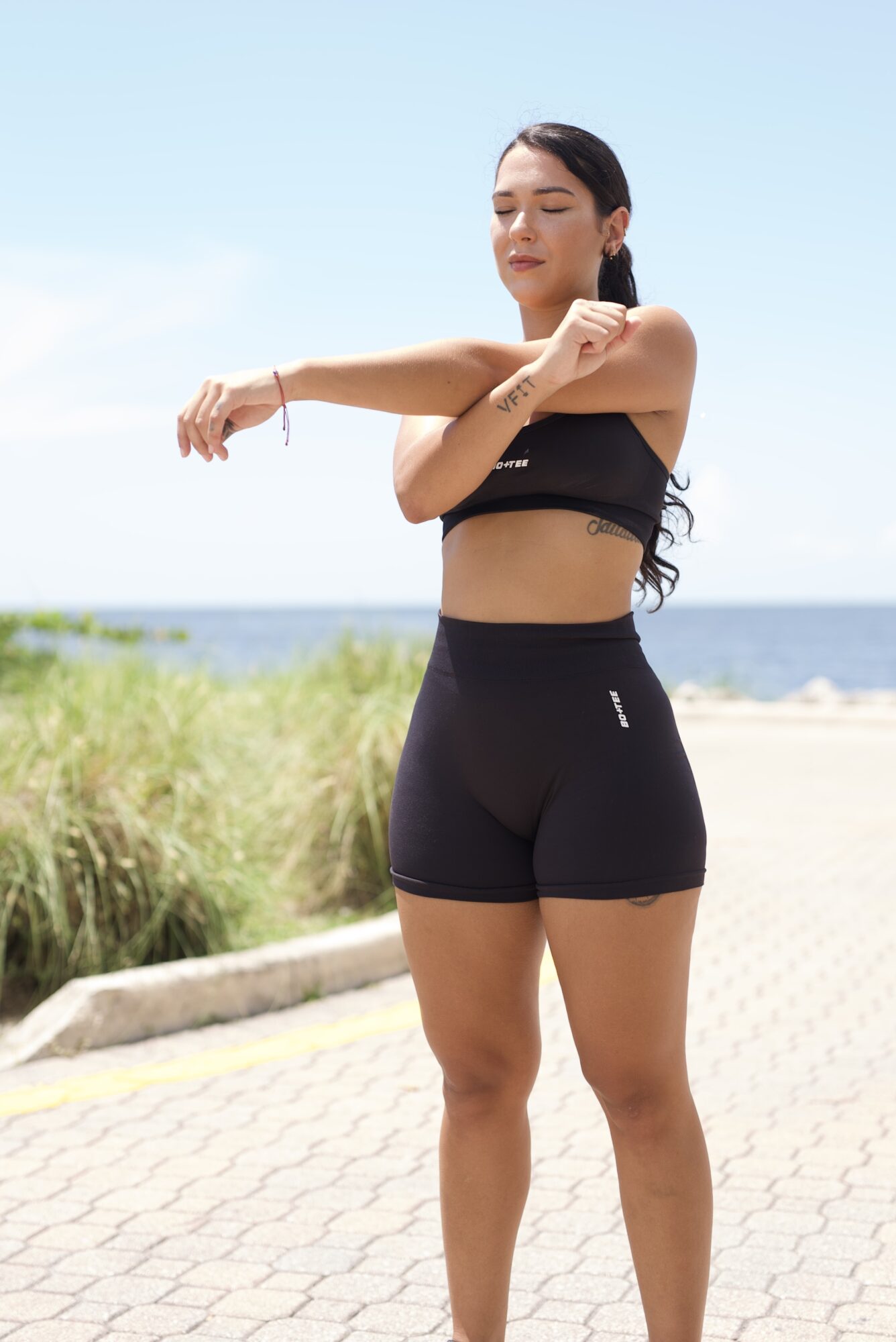 Woman stretching outdoors near beach, wearing black sports bra and shorts, with her eyes closed and arms extended.