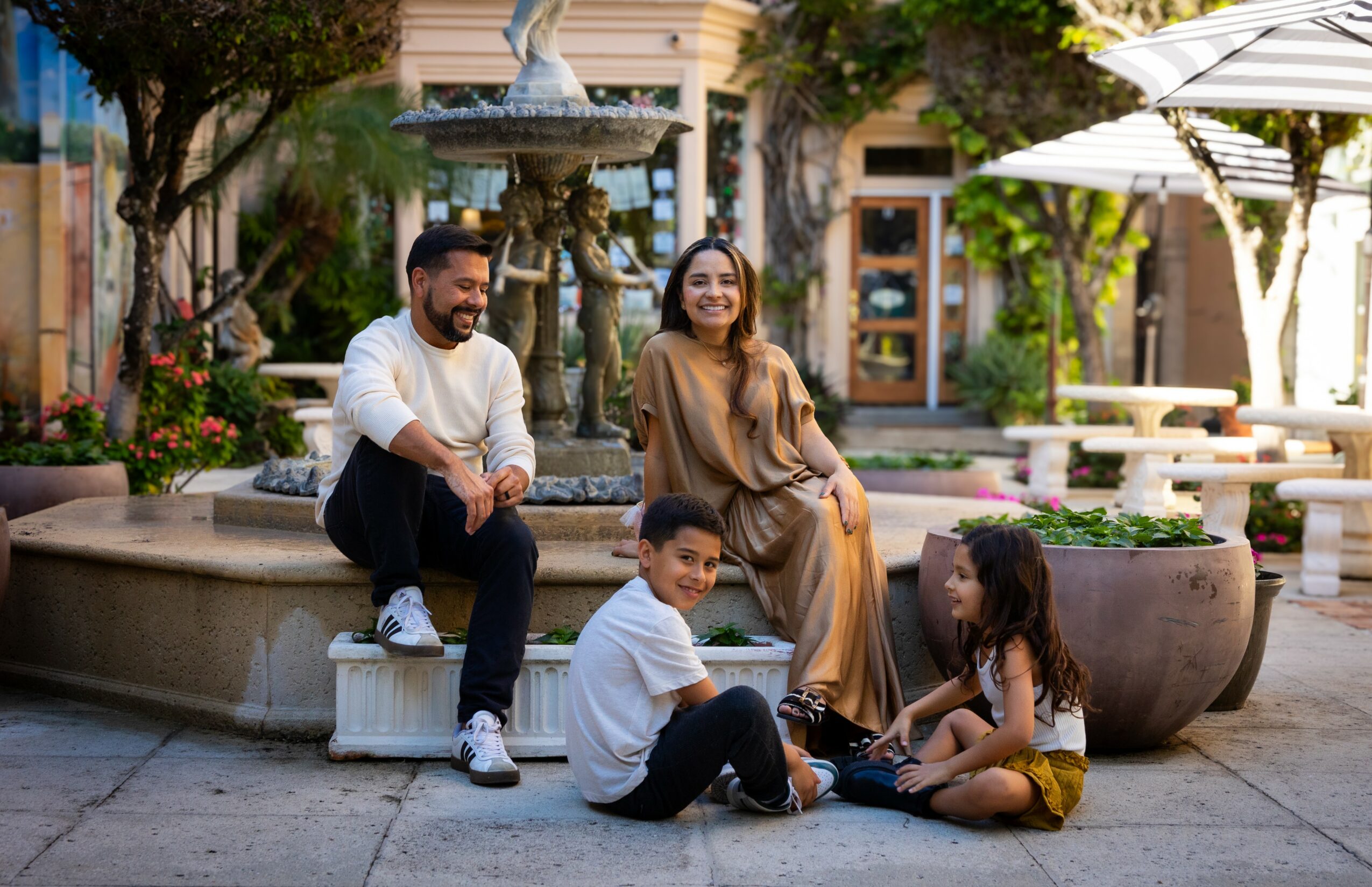 Four people sitting on a fountain in a courtyard with trees and buildings, smiling and relaxing outdoors.