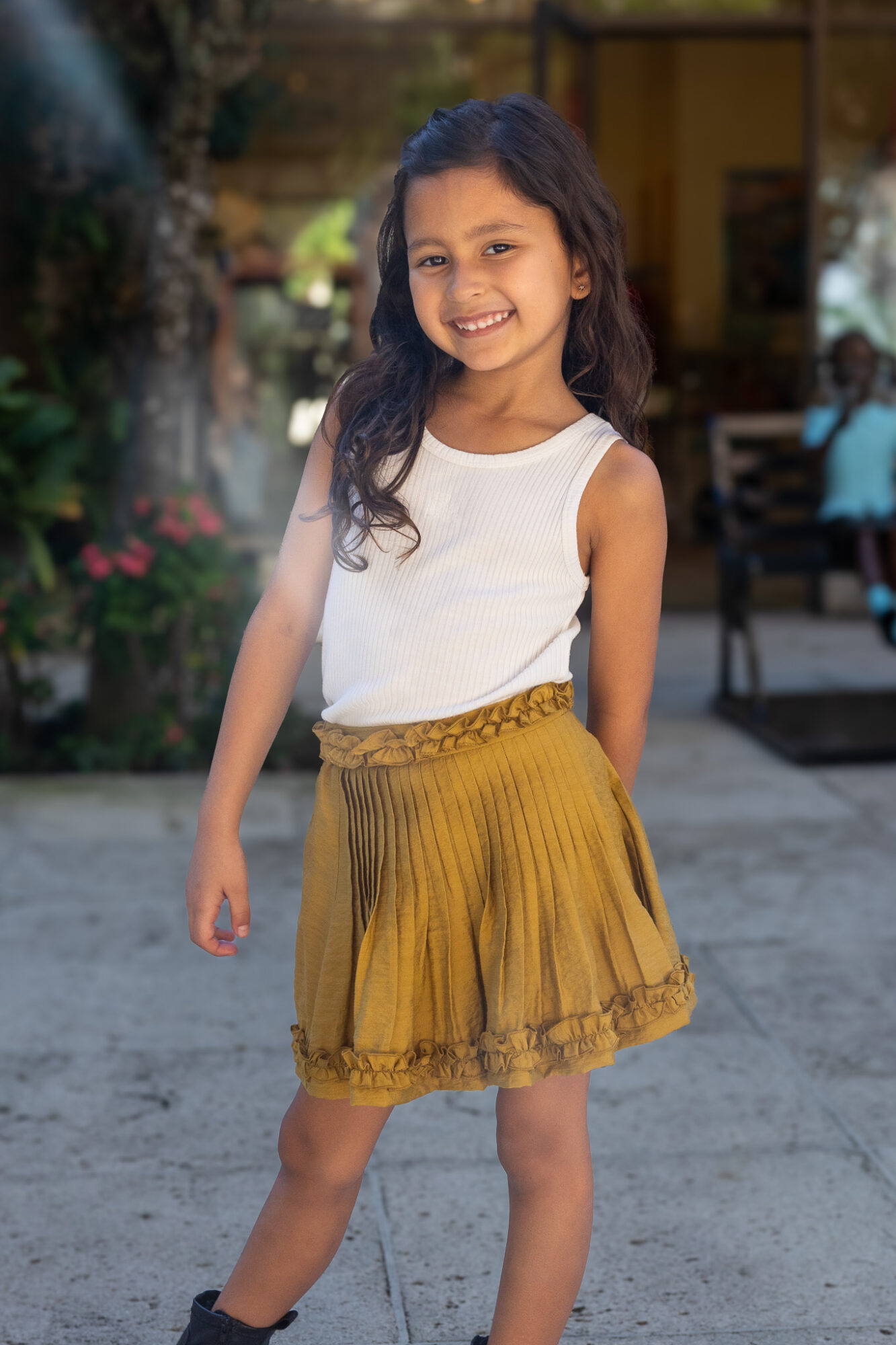 Young girl with long dark hair smiling, wearing a white sleeveless top and yellow skirt, standing outdoors.