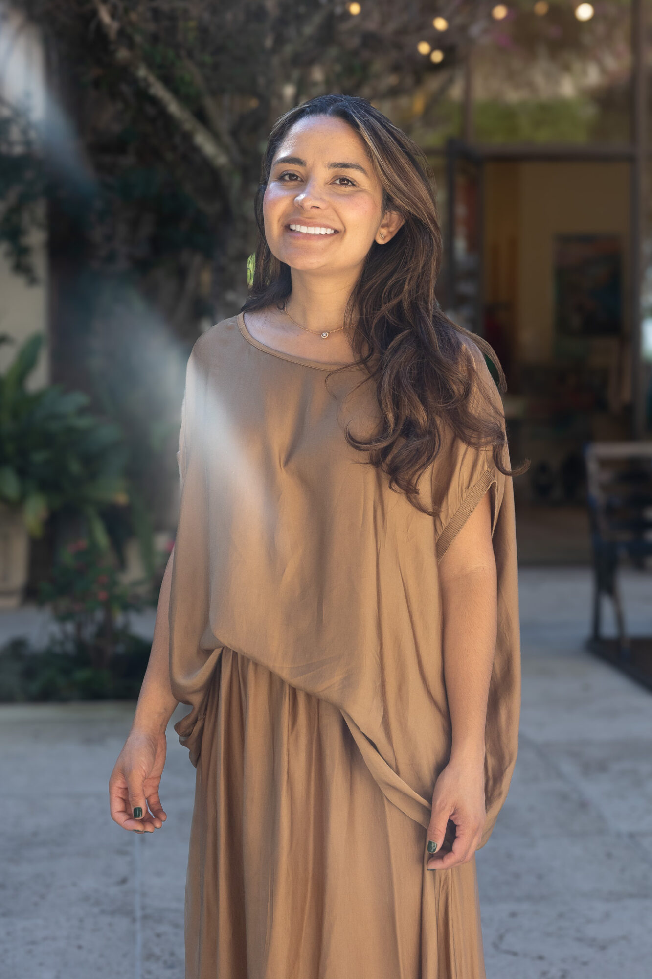 Smiling woman with long hair in a beige dress outdoors, trees and a building in background.