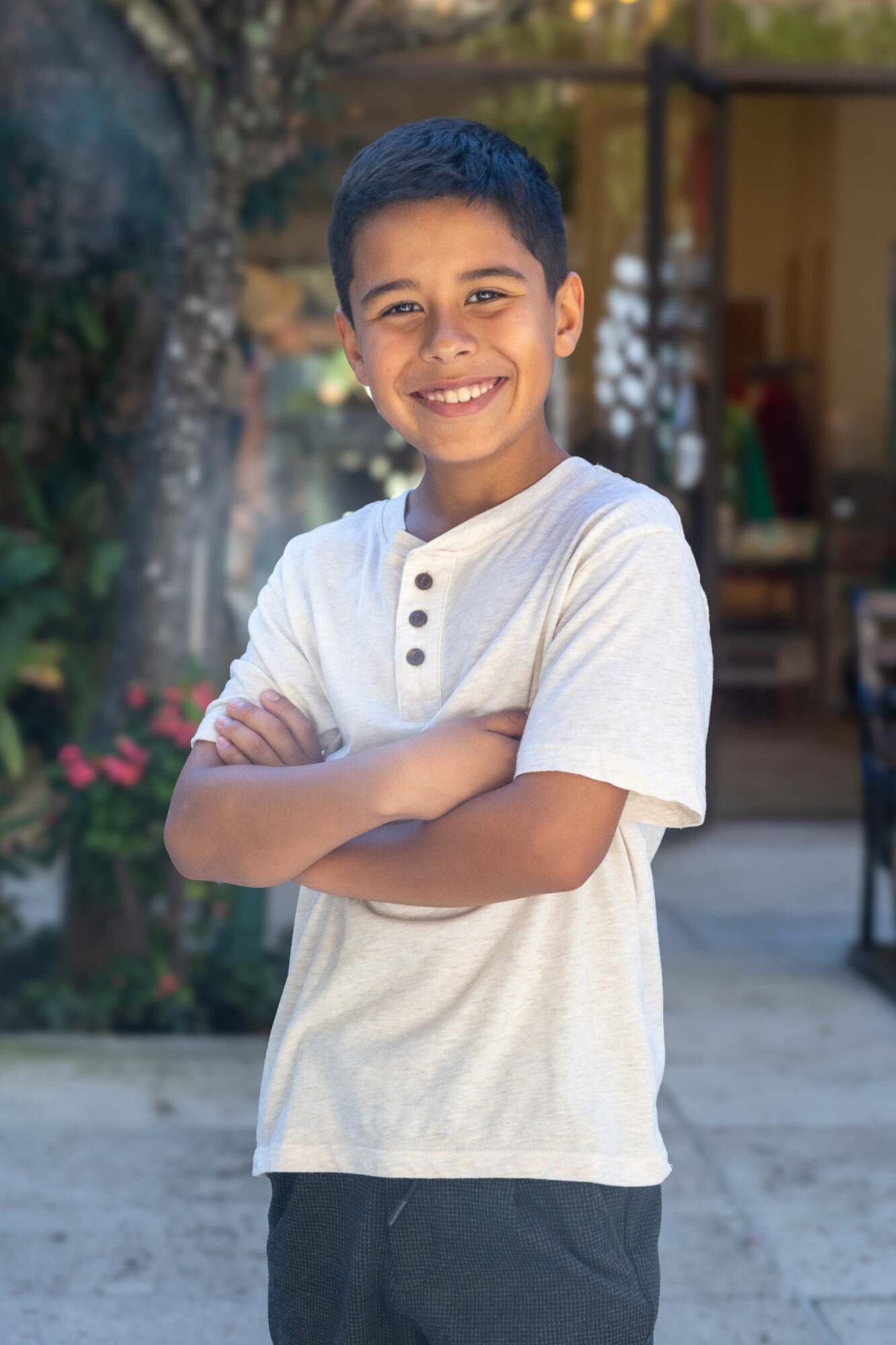 Smiling boy with arms crossed outdoors, wearing a white shirt and dark pants, in a garden setting.