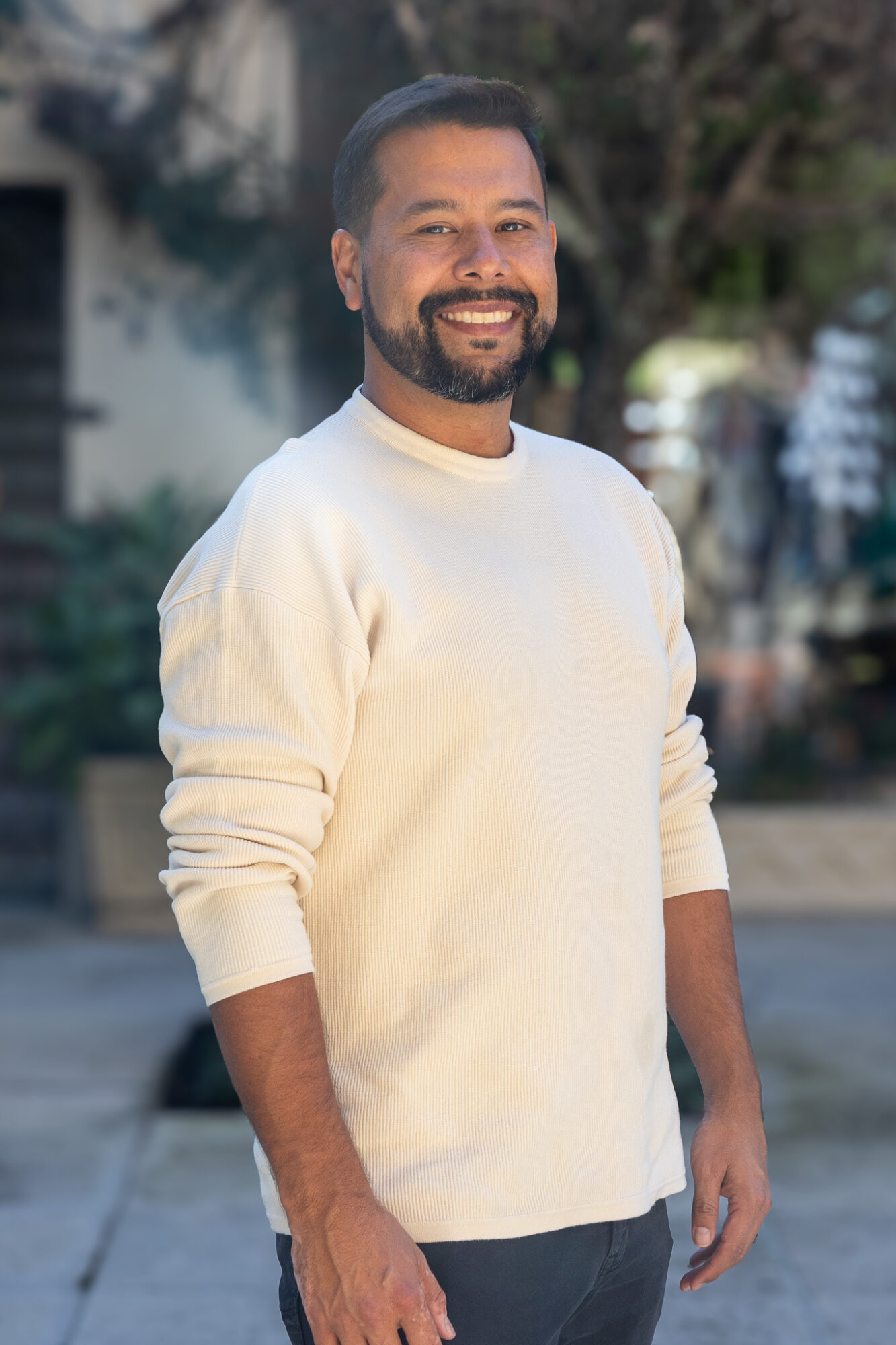 Smiling man with beard and mustache wearing a white sweater outdoors, background with trees and buildings.