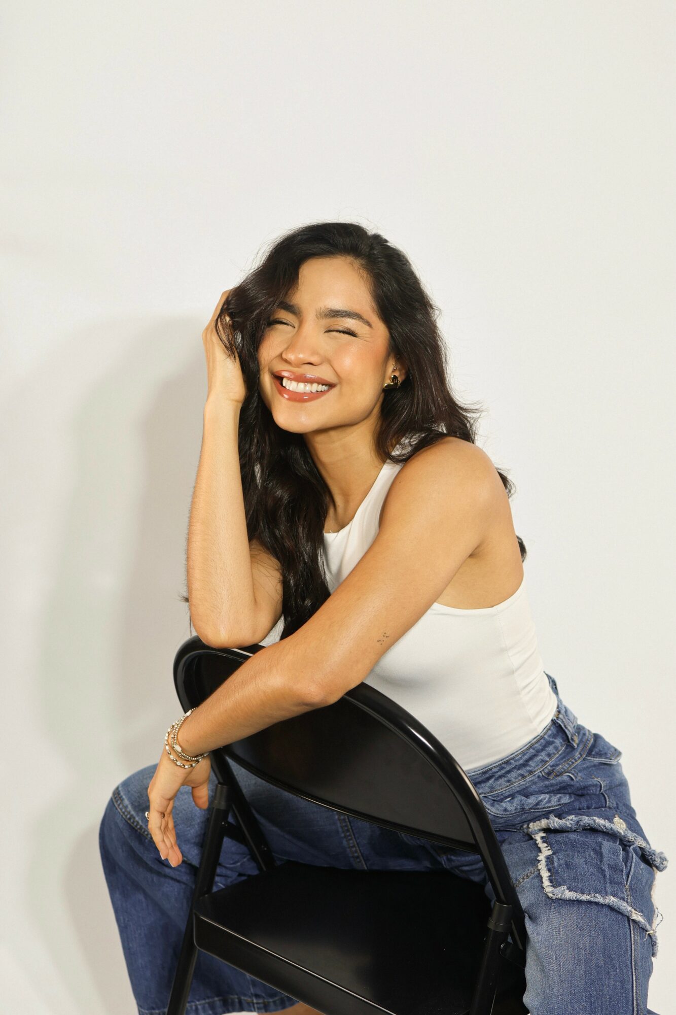 Smiling woman with long dark hair, wearing a white tank top and jeans, sitting on a black chair against a plain background.