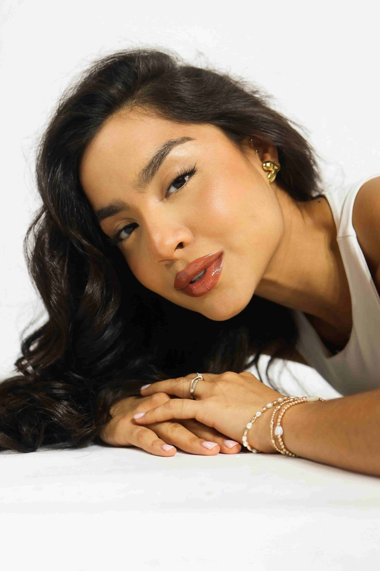 Woman with long dark hair lying on her side, resting her head on her hands, smiling softly, wearing jewelry and a white top.