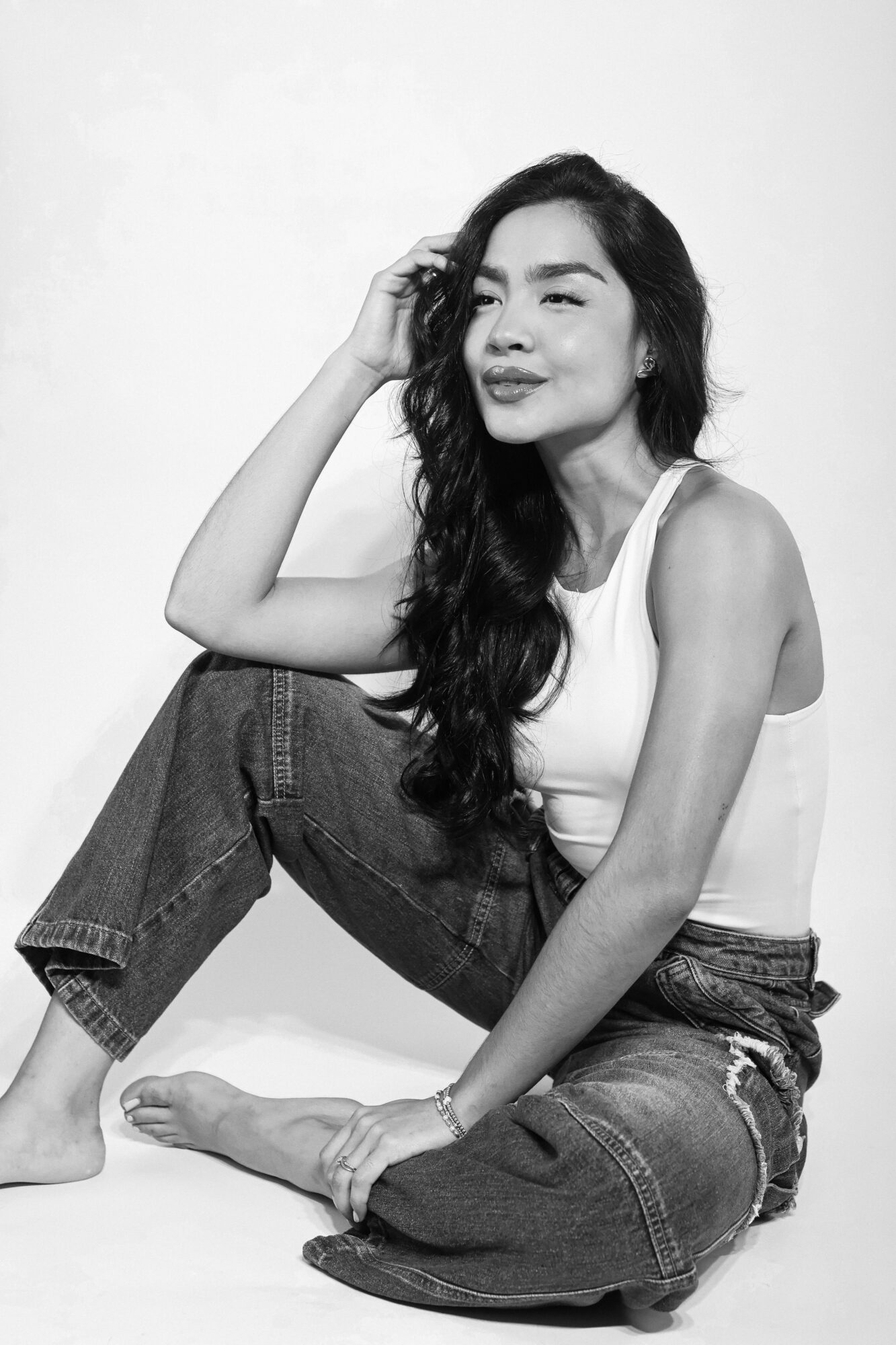 Woman with long wavy hair sitting on the floor, smiling, wearing a sleeveless top and jeans, against a plain background.