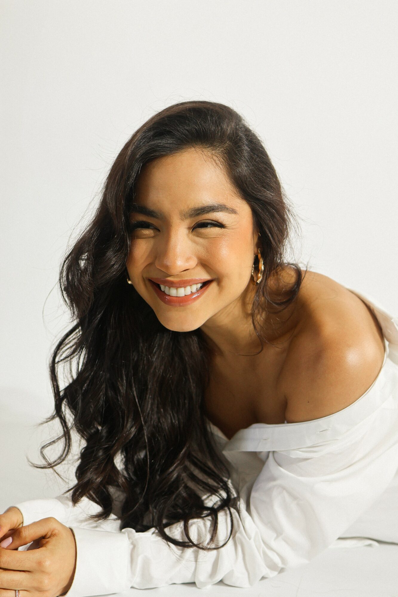 Smiling woman with long dark hair, wearing a white off-shoulder top, looking at the camera against a plain background.