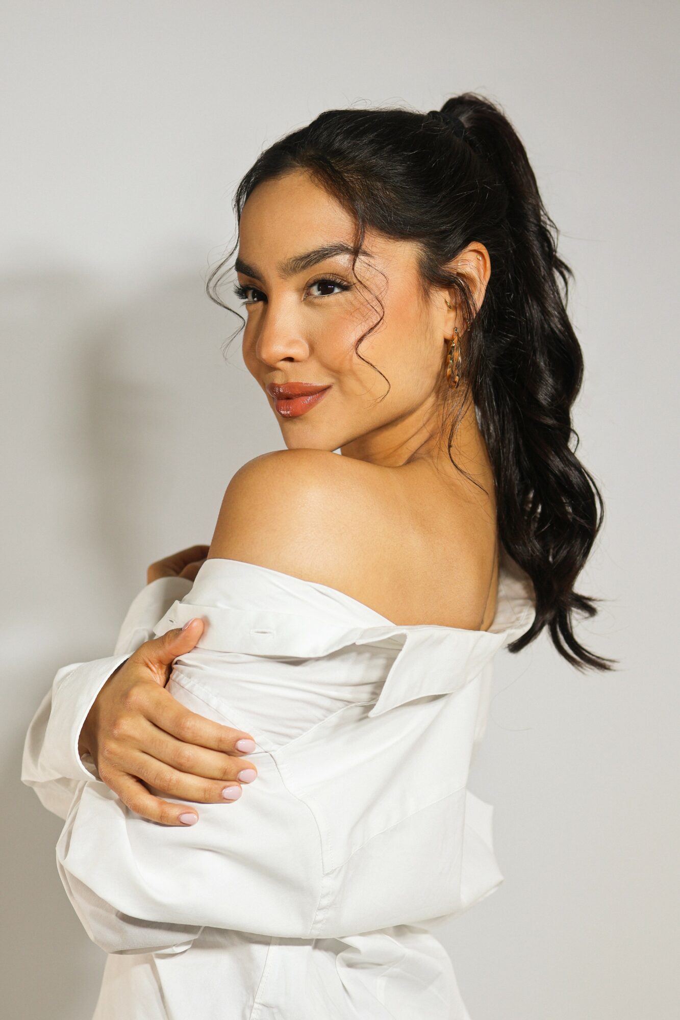 Young woman with long dark hair in a ponytail, wearing an off-shoulder white shirt, smiling softly, arms crossed.