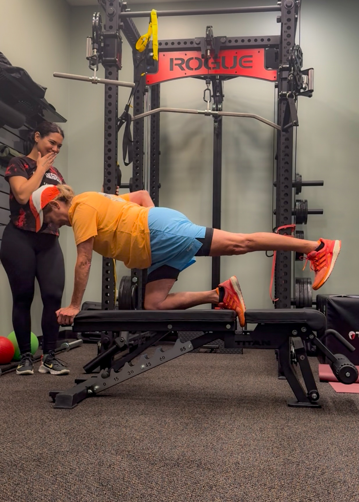 Person performing a plank exercise with one leg raised on a workout bench, in a gym setting with a trainer nearby.
