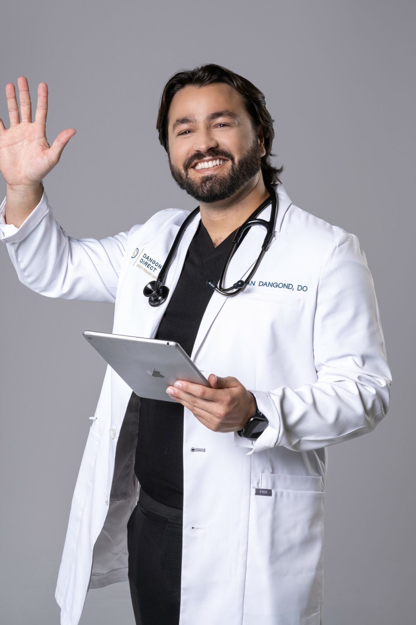 Male doctor with a beard in a white coat, holding a tablet, smiling, waving, against a gray background.