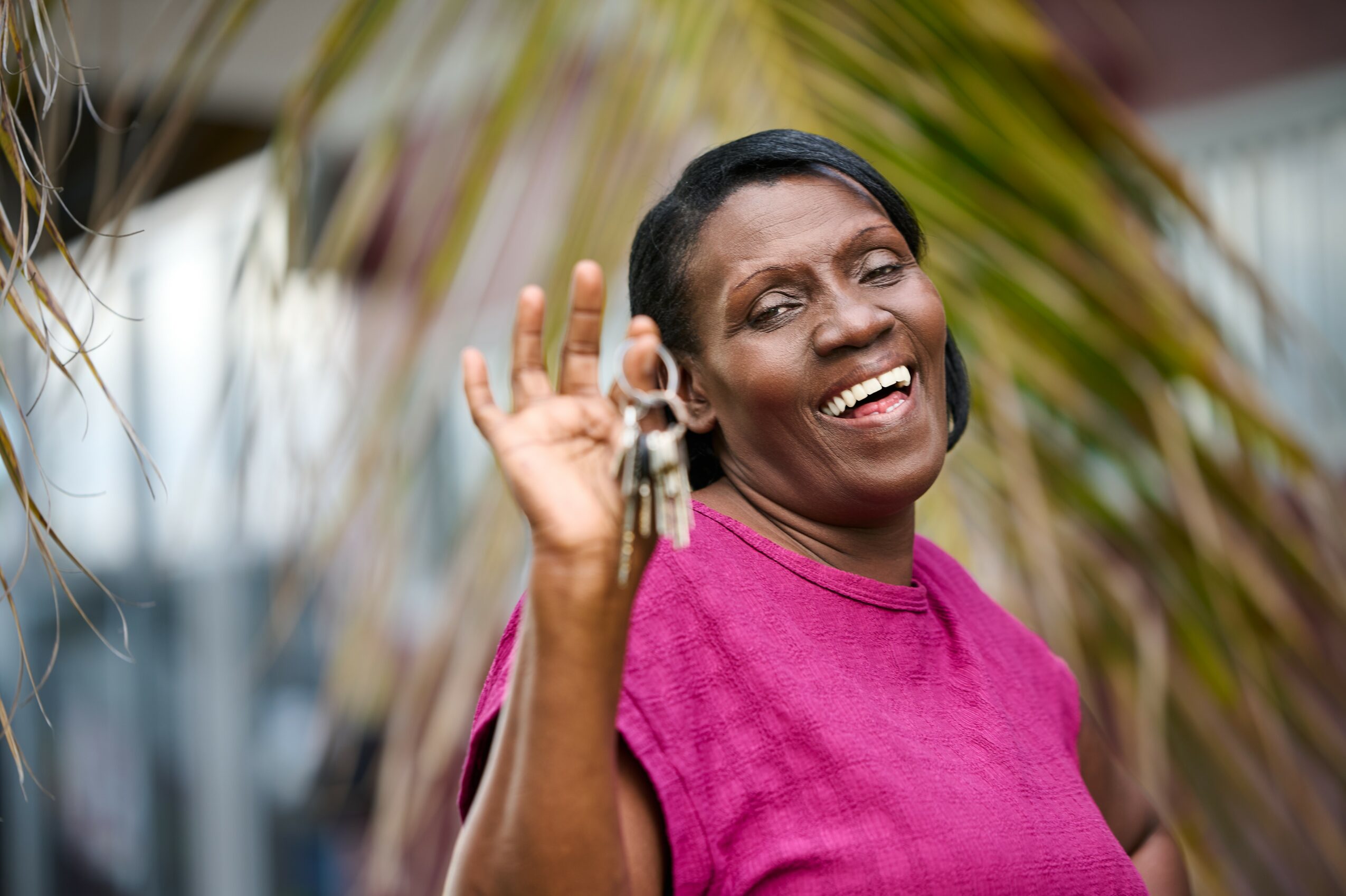 Smiling woman waving, wearing a pink top, outdoors with palm leaves in background.